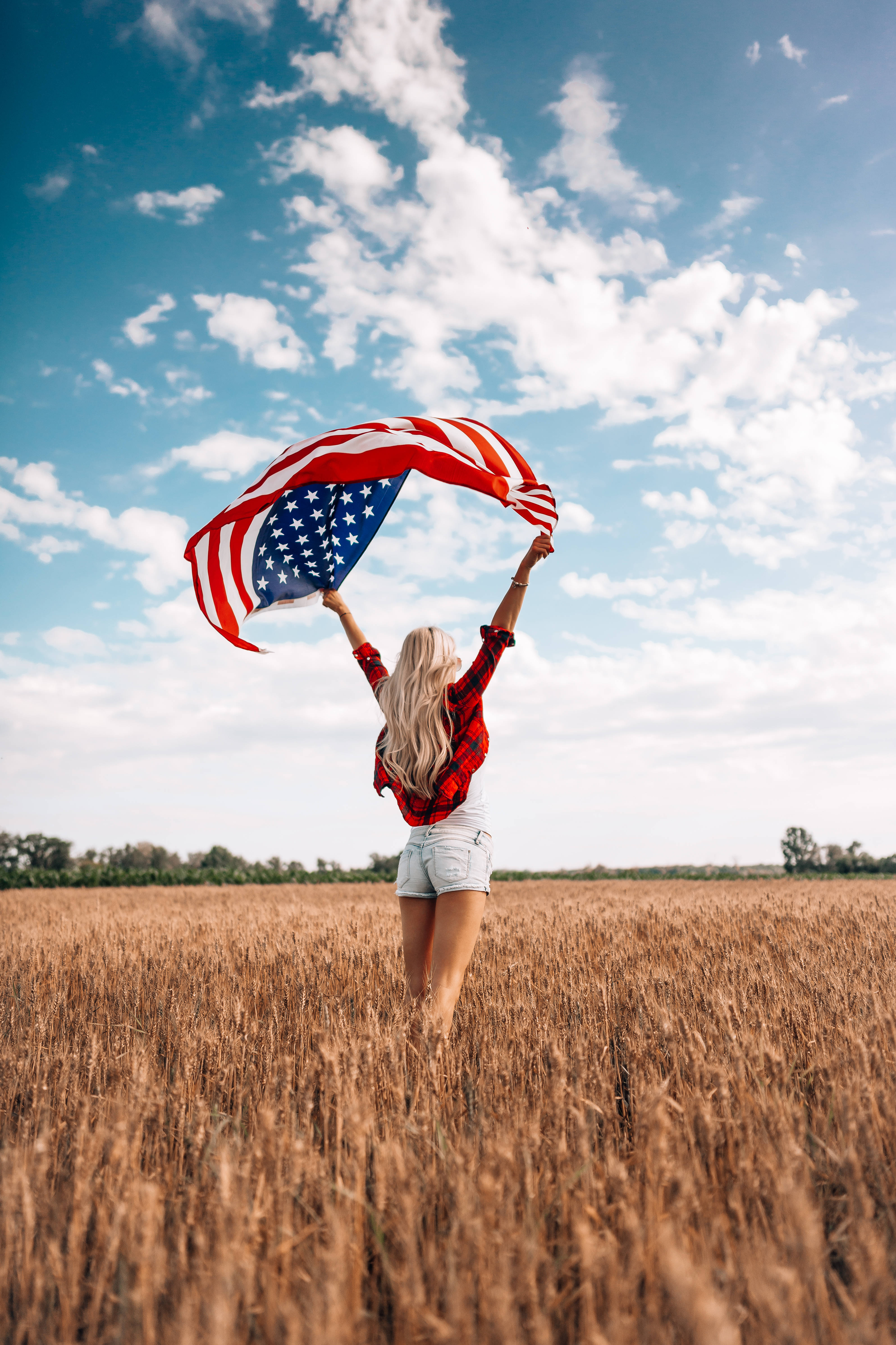 USA flag iPhone 桌布 壁紙 — 一位金髮女子站在金色麥田裡，高舉美國國旗，背景是晴朗的藍天和蓬鬆的白雲。