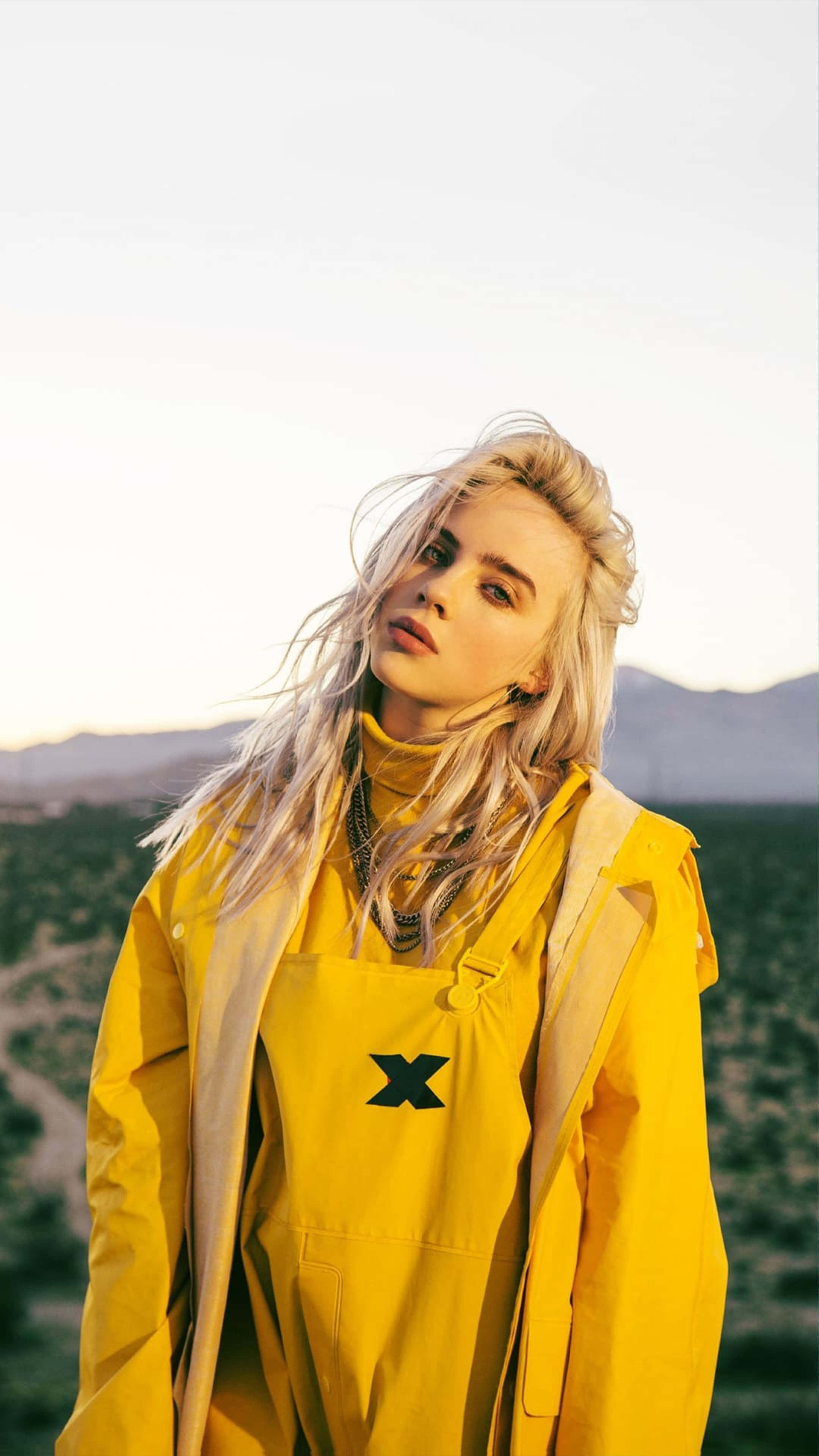 Yellow Raincoat 桌布 壁紙 — 一位金髮女子身穿亮黃色服裝，在柔和金色天空下，以沙漠和山脈為背景站立。