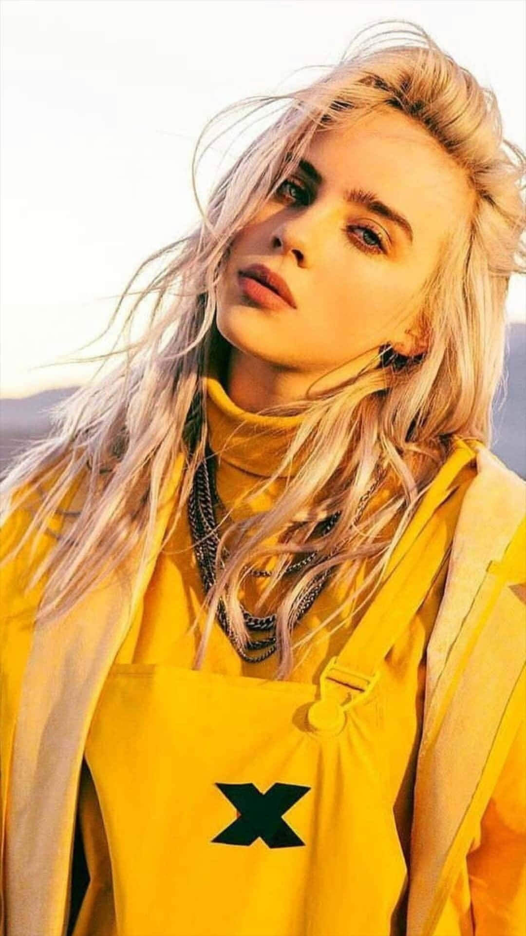 Billie Eilish 2021 桌布 壁紙 — 一位金髮女子身穿亮黃色服裝，頭髮被風吹起，在朦朧的戶外背景下向上凝視。