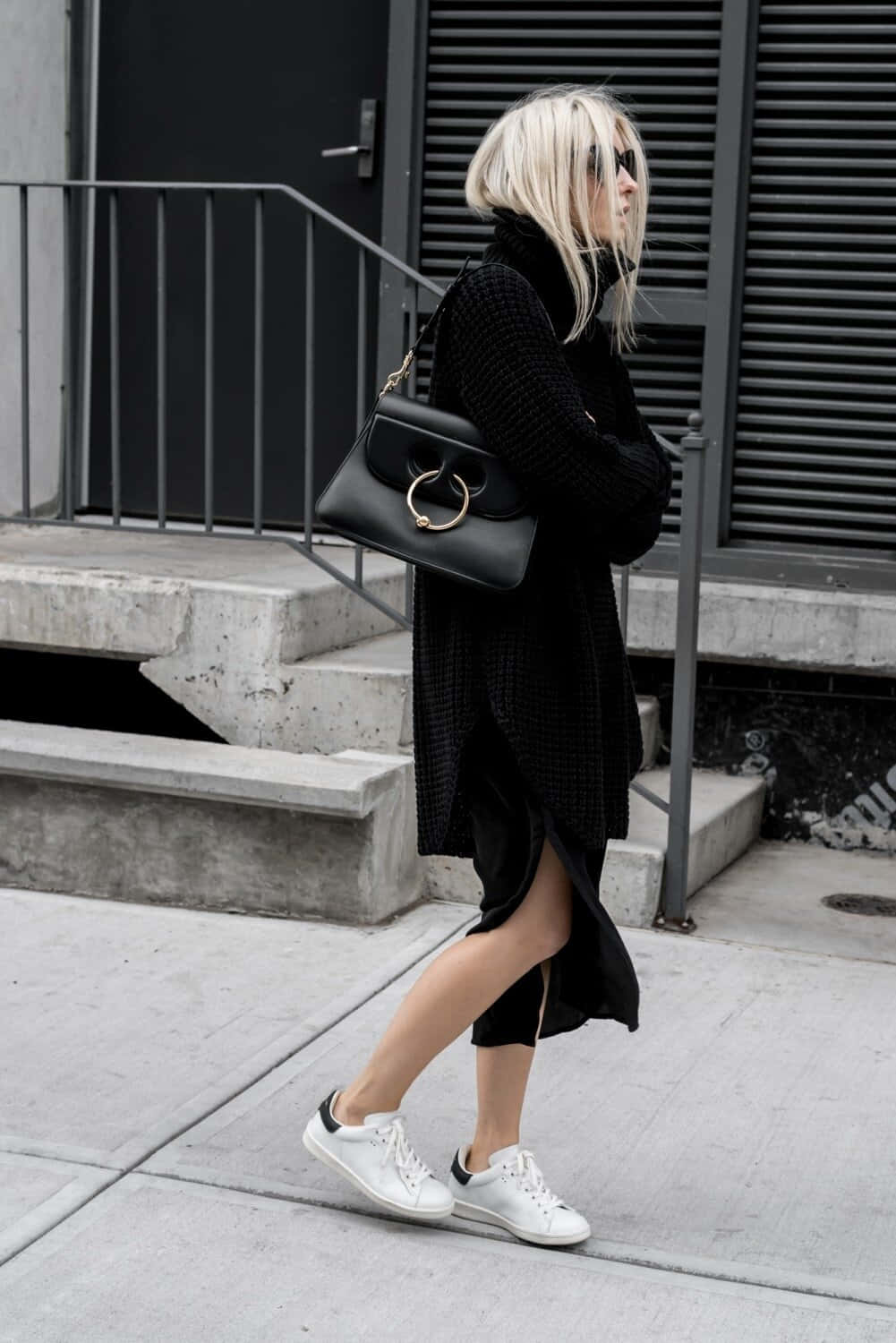Minimalist Fashion 桌布 壁紙 — 一位金髮女子身穿全黑衣服，走過混凝土人行道，經過金屬樓梯和深色建築物外牆。