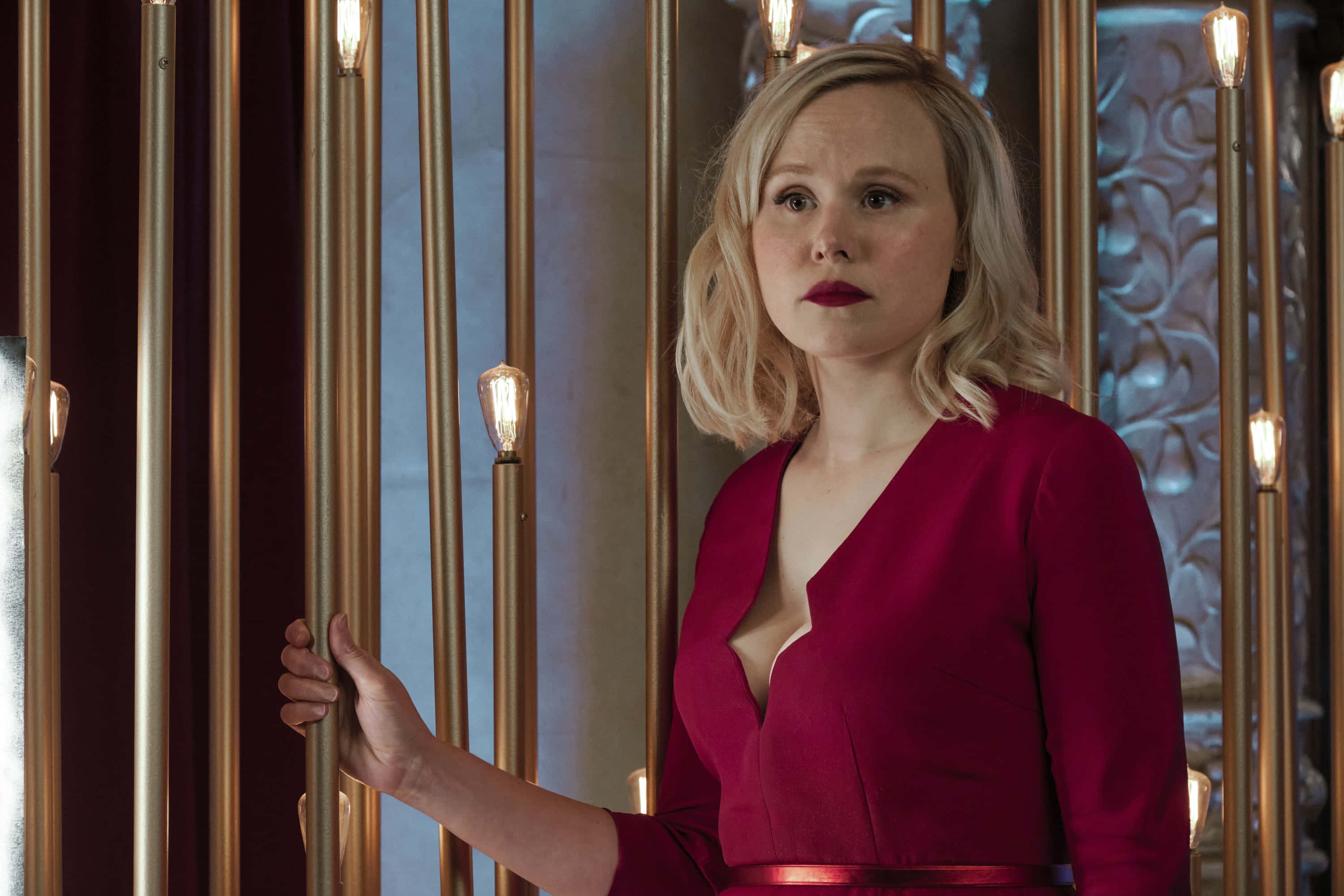 Alison Pill 桌布 壁紙 — 一位金髮女子身穿深紅色連衣裙，站在豪華室內發光的金色垂直管之間。
