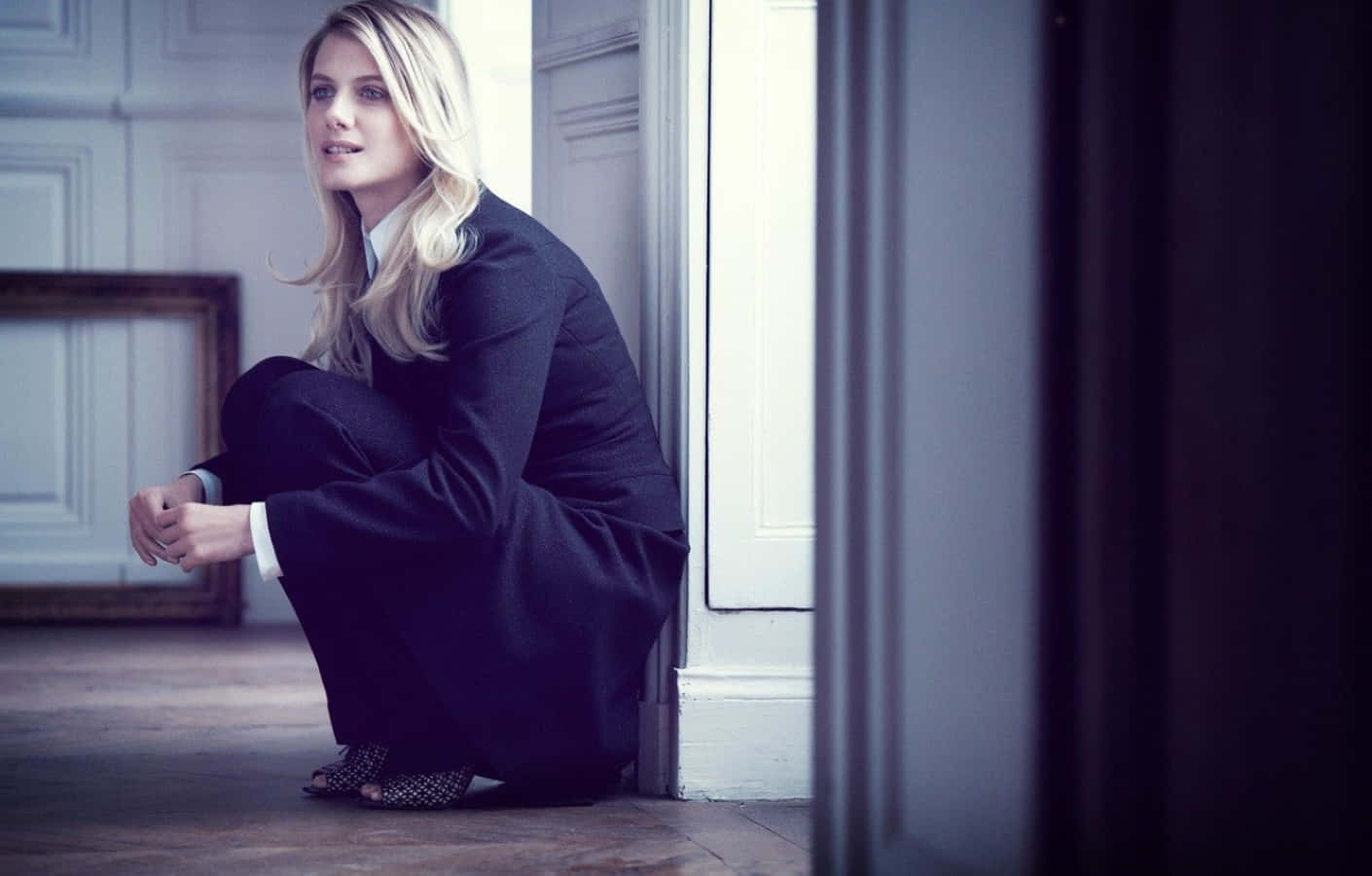 Mélanie Laurent 桌布 壁紙 — 一位金髮女子身穿深色西裝，蹲在門口旁，房間光線柔和，配有經典鑲板和木地板。