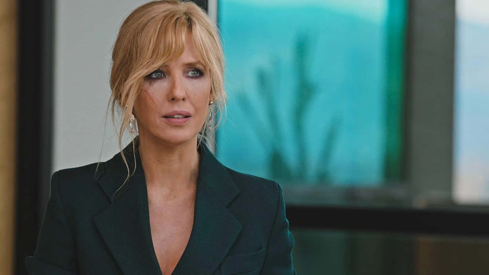 Kelly Reilly 桌布 壁紙 — 一位金髮女子身穿深色西裝外套，臉頰有瘀傷，在模糊的藍綠色窗戶背景下專注地凝視。