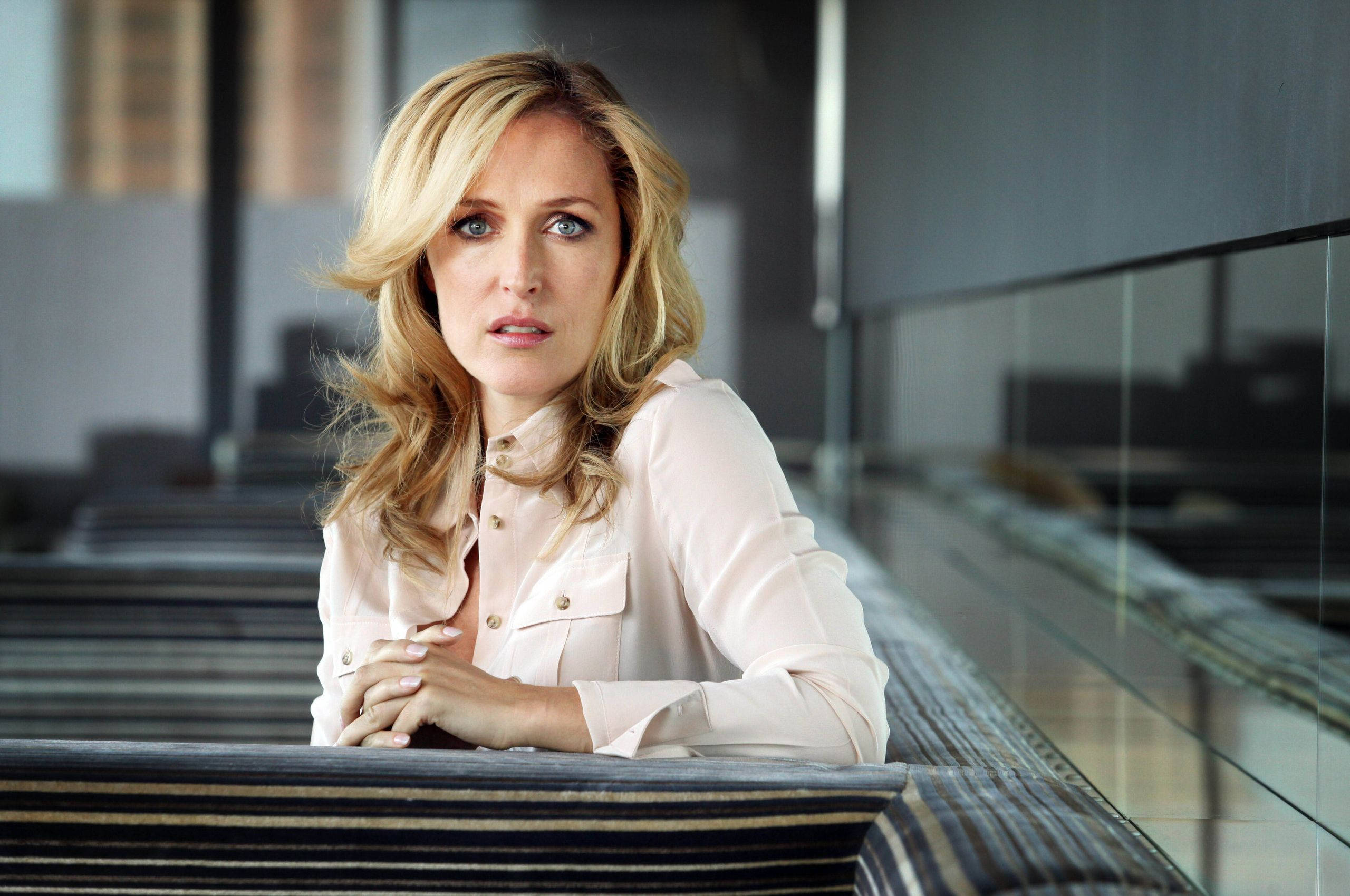 gillian anderson 桌布 壁紙 — 一位金髮女子身穿淺粉紅色襯衫，在現代玻璃牆環境中，表情嚴肅地靠在條紋欄杆上。