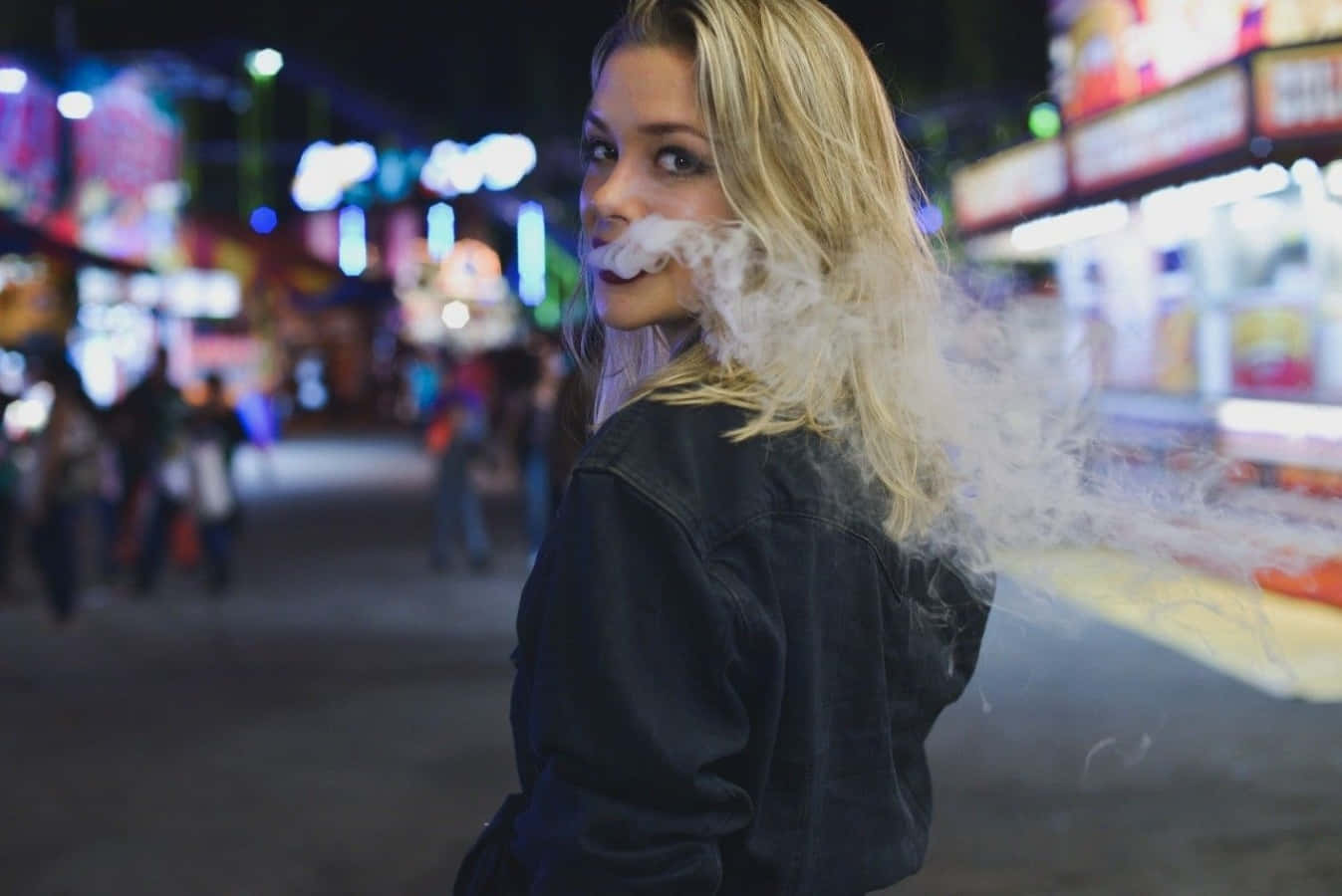 抽菸 桌布 壁紙 — 一位金髮女子身穿牛仔外套，在夜晚霓虹燈閃爍的街道上回頭呼出煙霧。