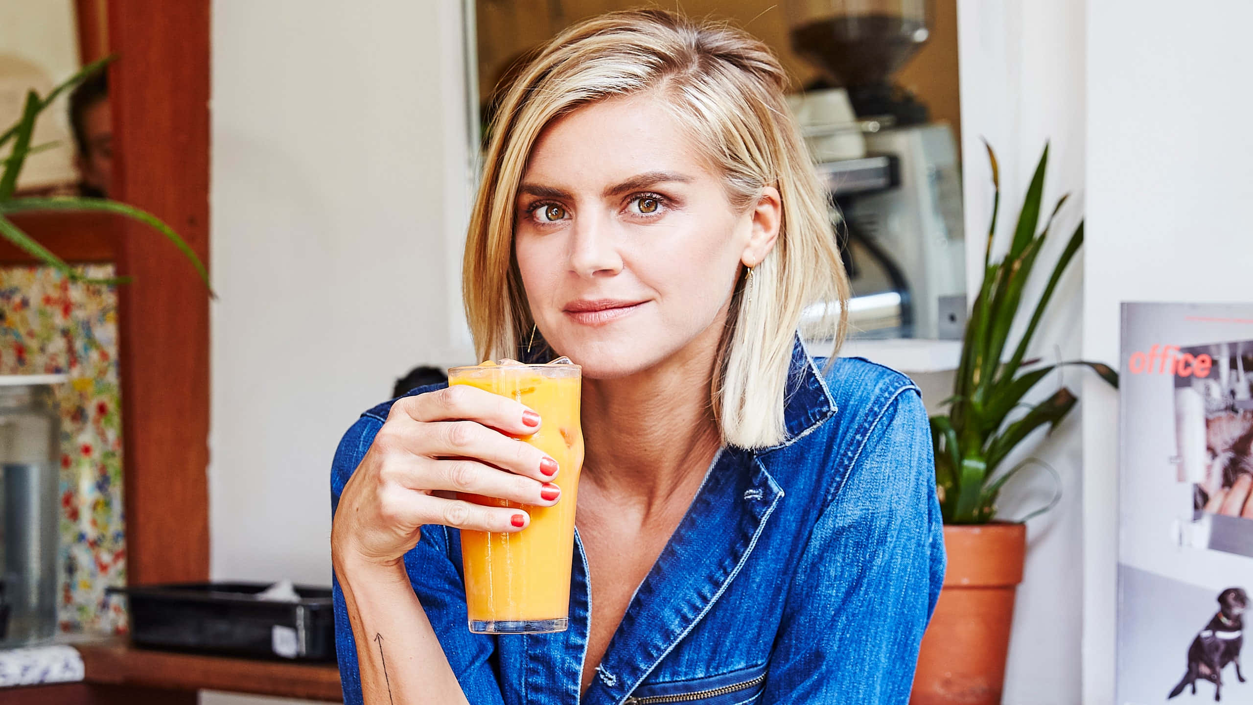eliza coupe 桌布 壁紙 — 一位金髮女子身穿牛仔外套，在舒適的咖啡廳環境中手持一杯柳橙汁，對著鏡頭微笑。