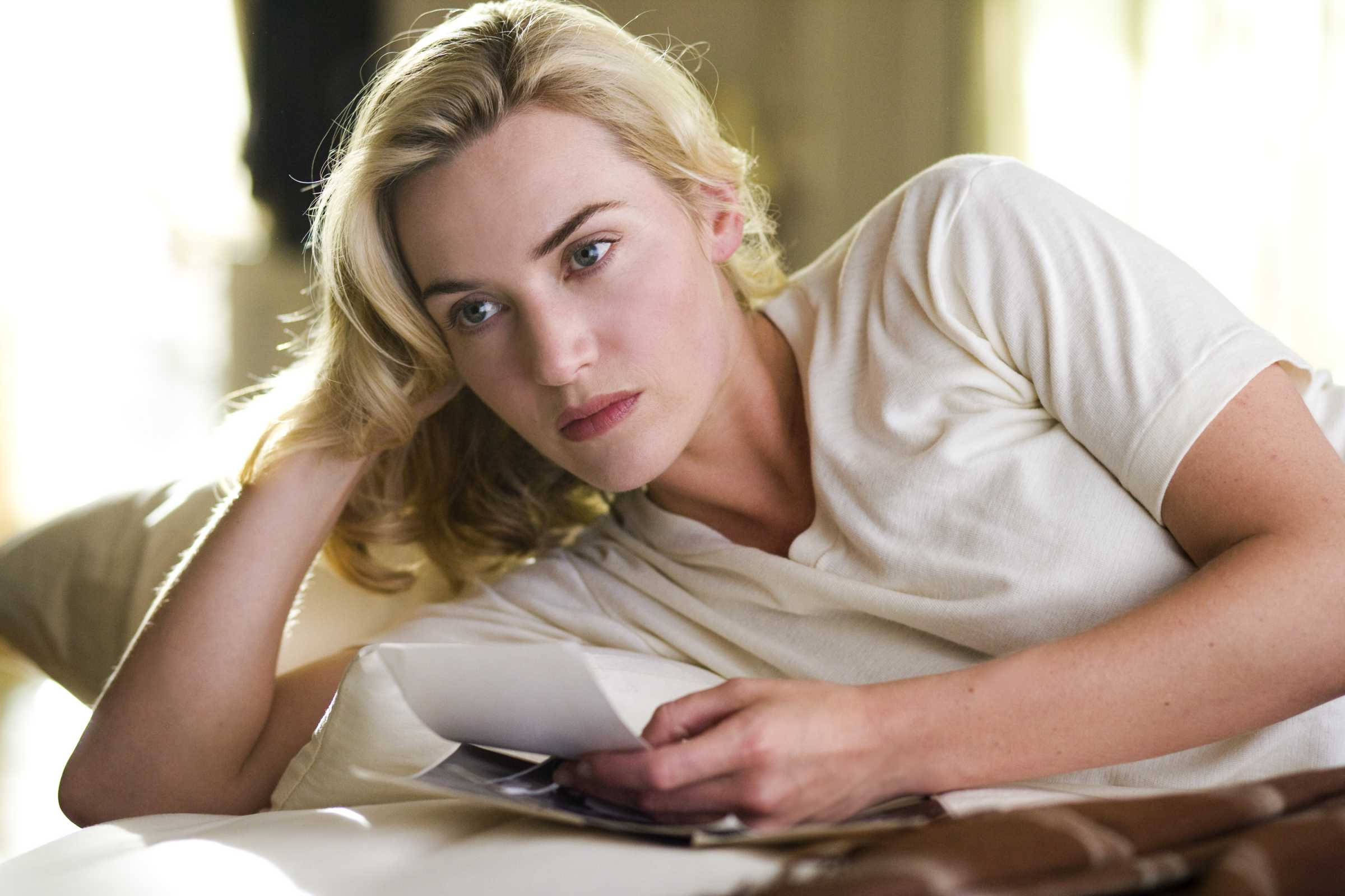 Kate Winslet 桌布 壁紙 — 一位金髮女子身穿白色T恤躺在床上，手持文件，表情嚴肅，陽光照射進來。
