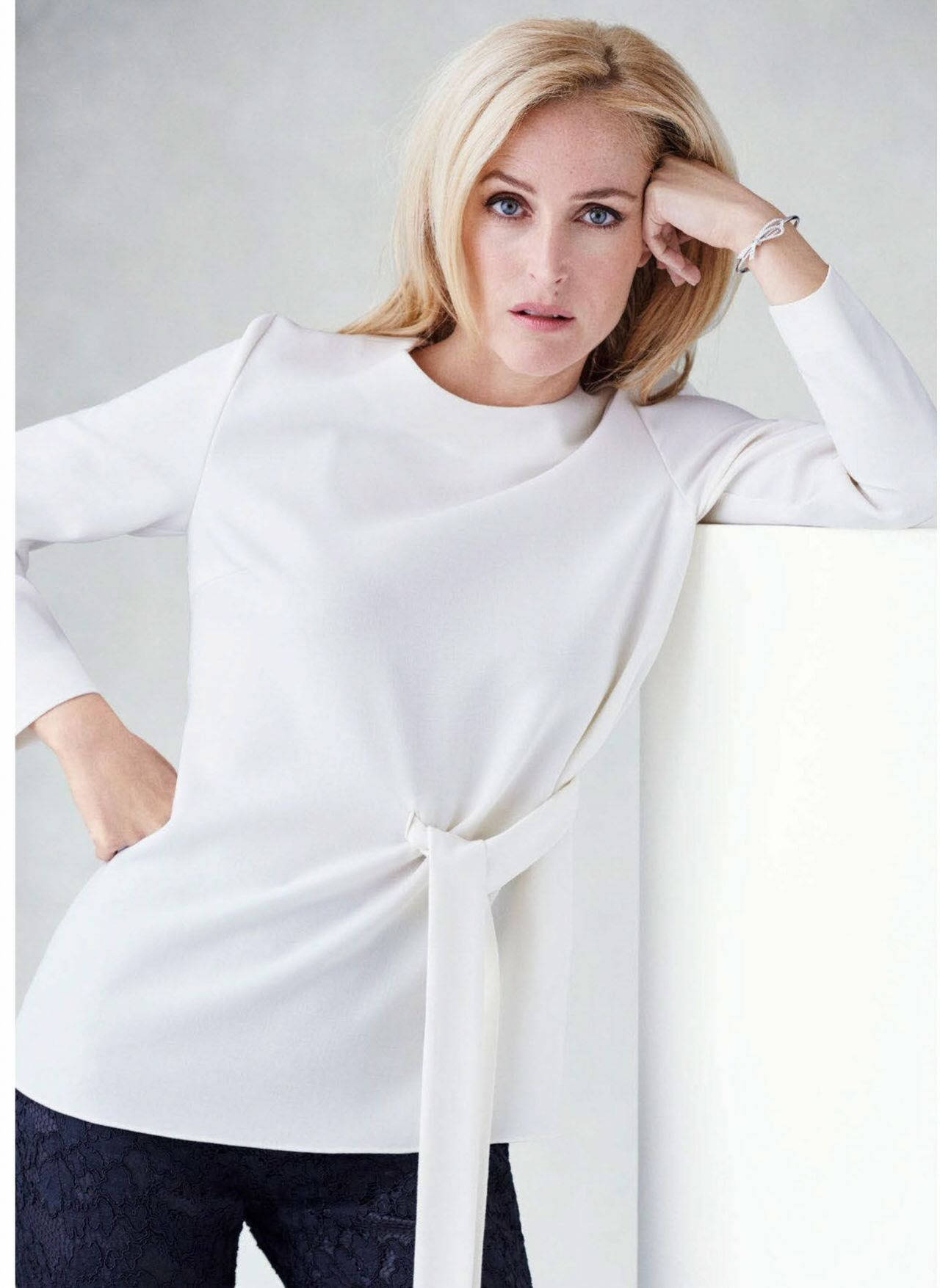 gillian anderson 桌布 壁紙 — 一位金髮女子身穿白色垂褶襯衫，一手放在頭上，另一手插在口袋裡，靠著牆。