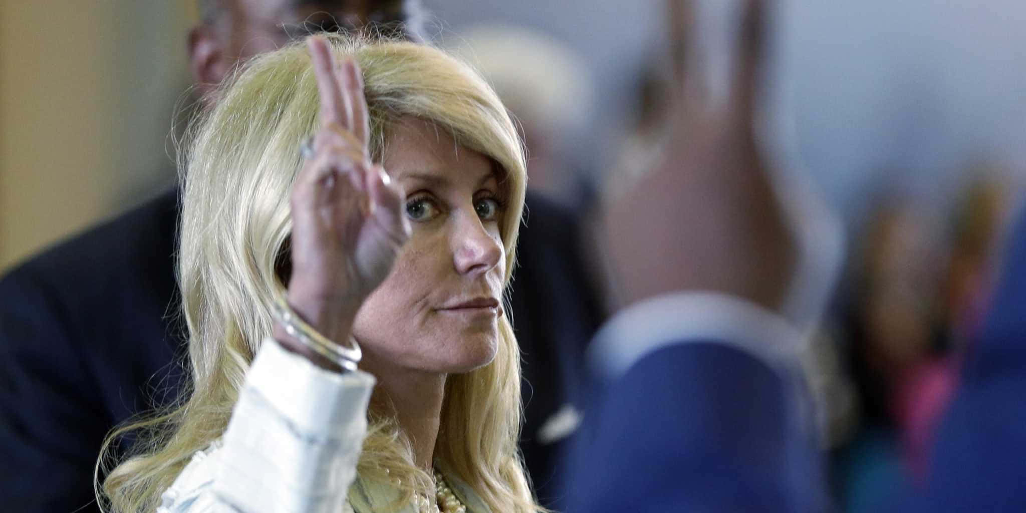 peace out 桌布 壁紙 — 一位金髮女子身穿白色外套，在正式室內環境中舉手，身後有模糊的人影。