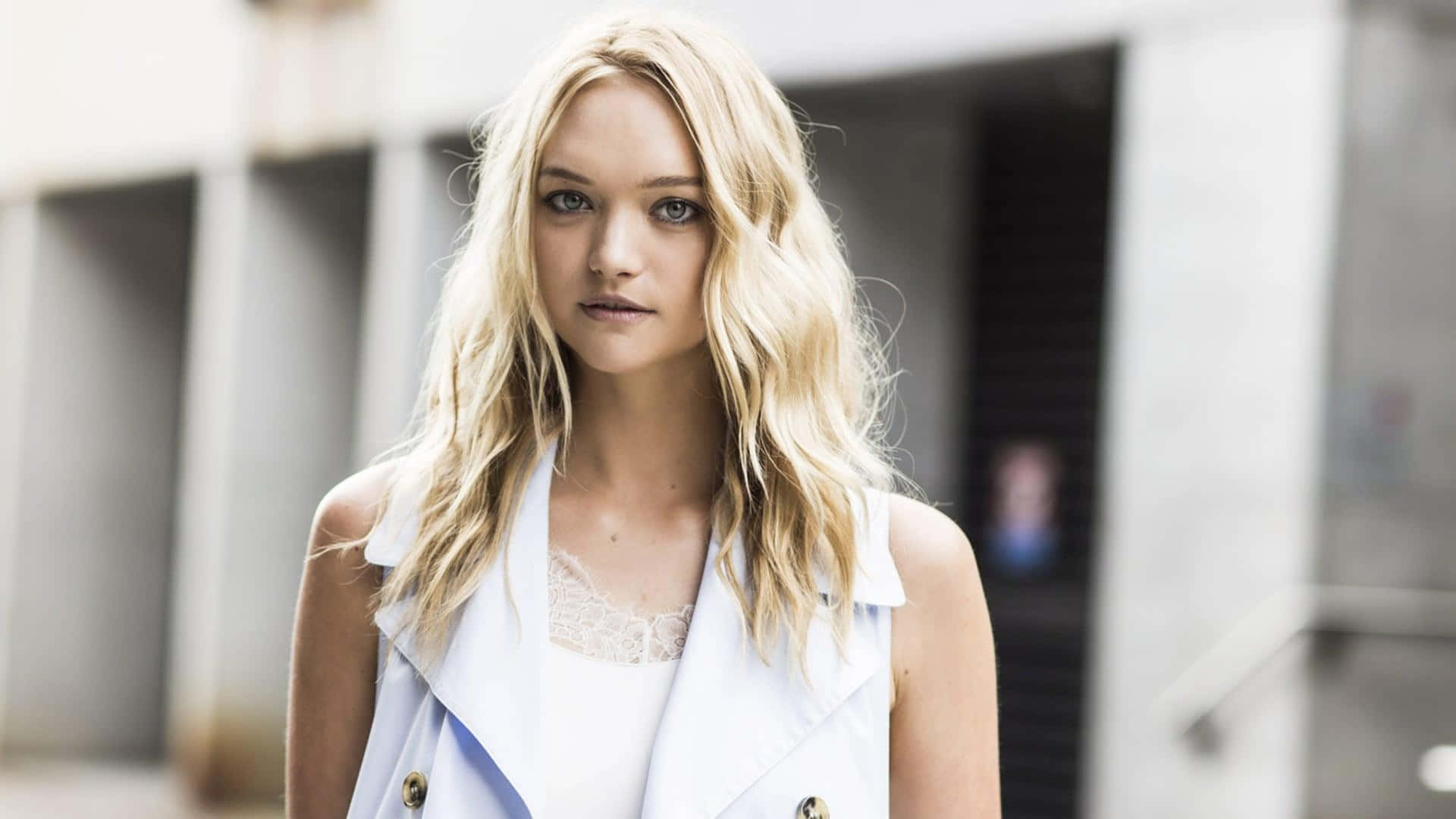 Gemma Ward 桌布 壁紙 — 一位金髮女子身穿白色無袖背心，自信地站在模糊的城市建築背景前。