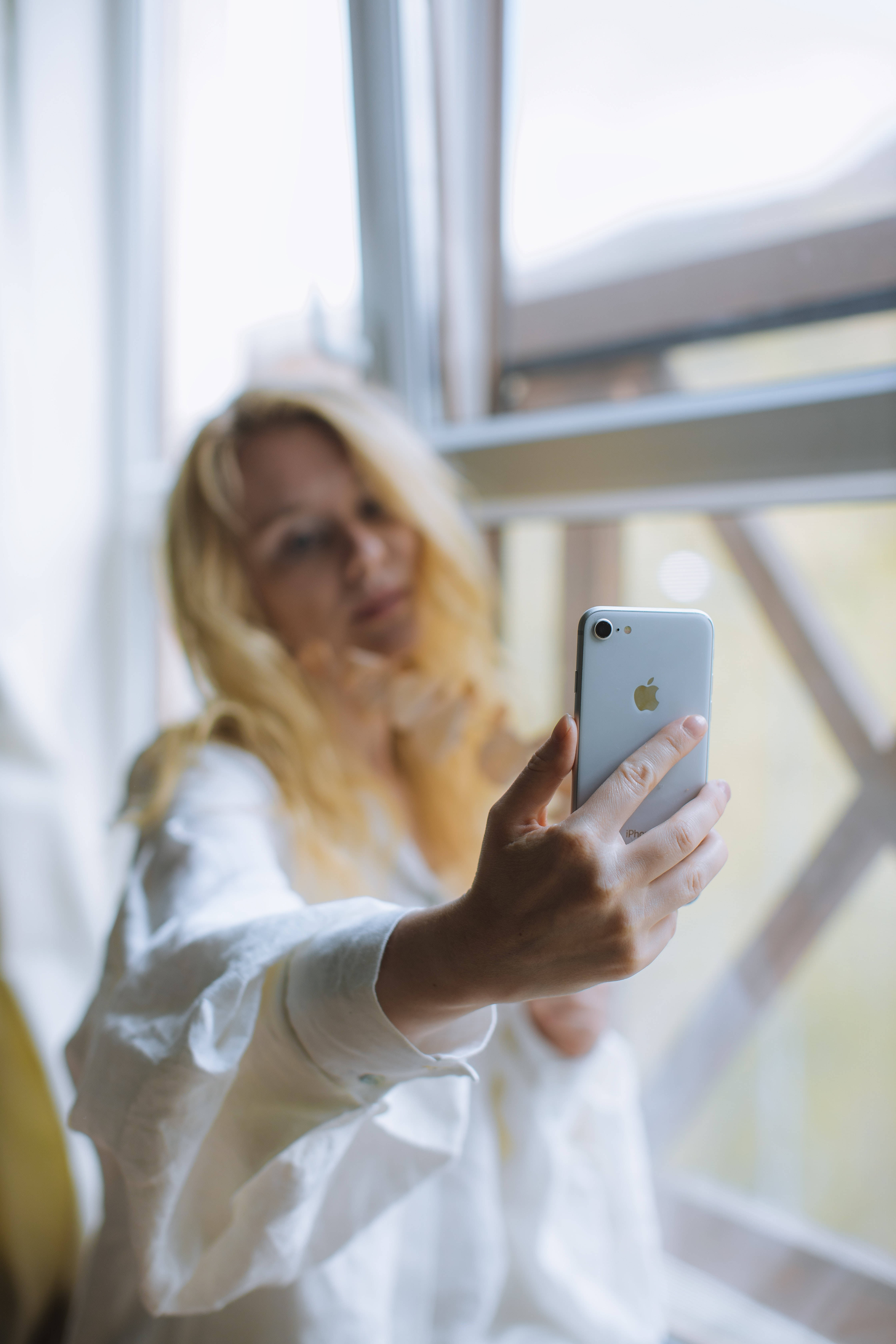 iPhone 桌布 壁紙 — 一位金髮女子身穿白色襯衫，在明亮的窗邊用銀色iPhone自拍。