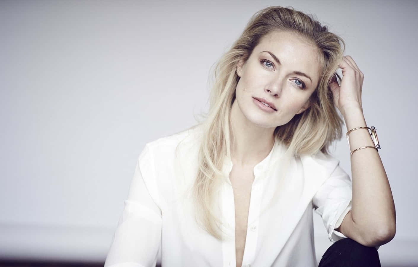 Mélanie Laurent 桌布 壁紙 — 一位金髮女子身穿白色襯衫，在柔和的灰色背景前若有所思地觸摸頭髮。