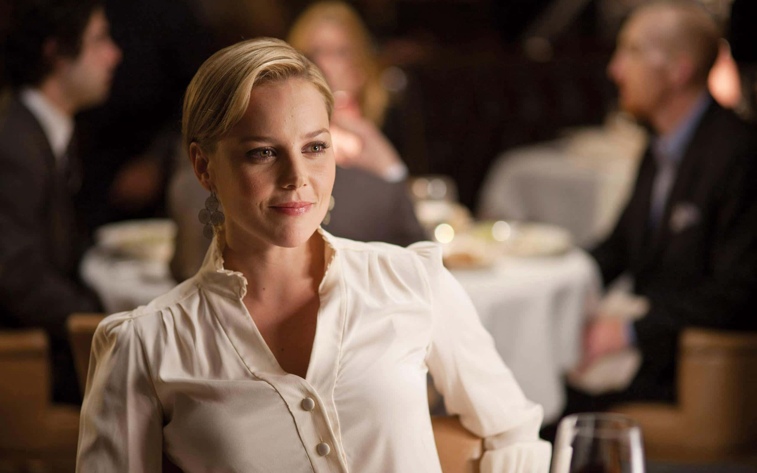 Abbie Cornish 桌布 壁紙 — 一位金髮女子身穿白色襯衫，在燈光昏暗的高級餐廳中微笑，背景有模糊的顧客。
