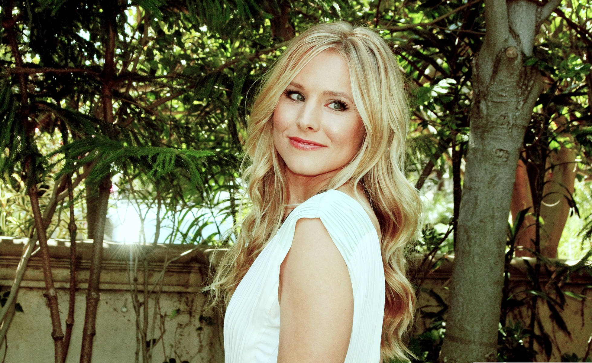 kristen bell 桌布 壁紙 — 一位金髮女子身穿白色連衣裙，在陽光穿過樹林的茂密綠葉中溫柔微笑。