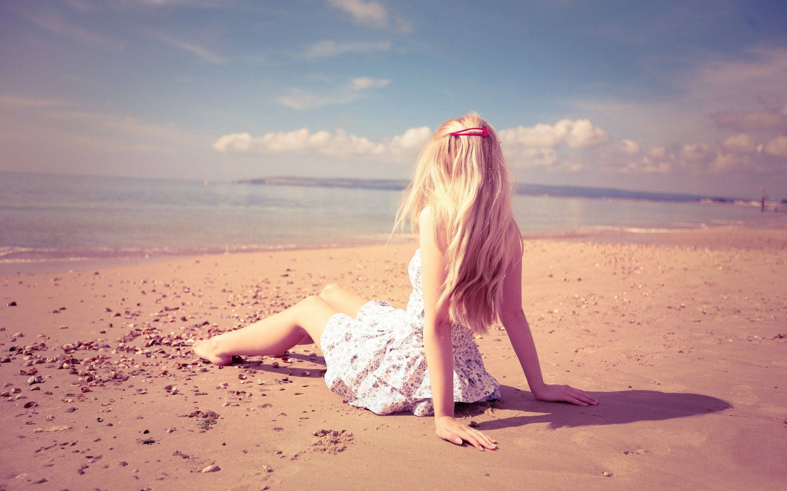 Beach Girl 桌布 壁紙 — 一位金髮女子身穿白色連衣裙，坐在陽光明媚的海灘上，凝視著部分多雲的天空下的海洋。