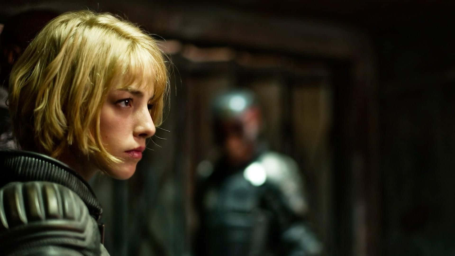 Olivia Thirlby 桌布 壁紙 — 一位金髮女子身穿盔甲，在昏暗的木造房間中，身後有一個模糊的盔甲身影，她專注地向前凝視。