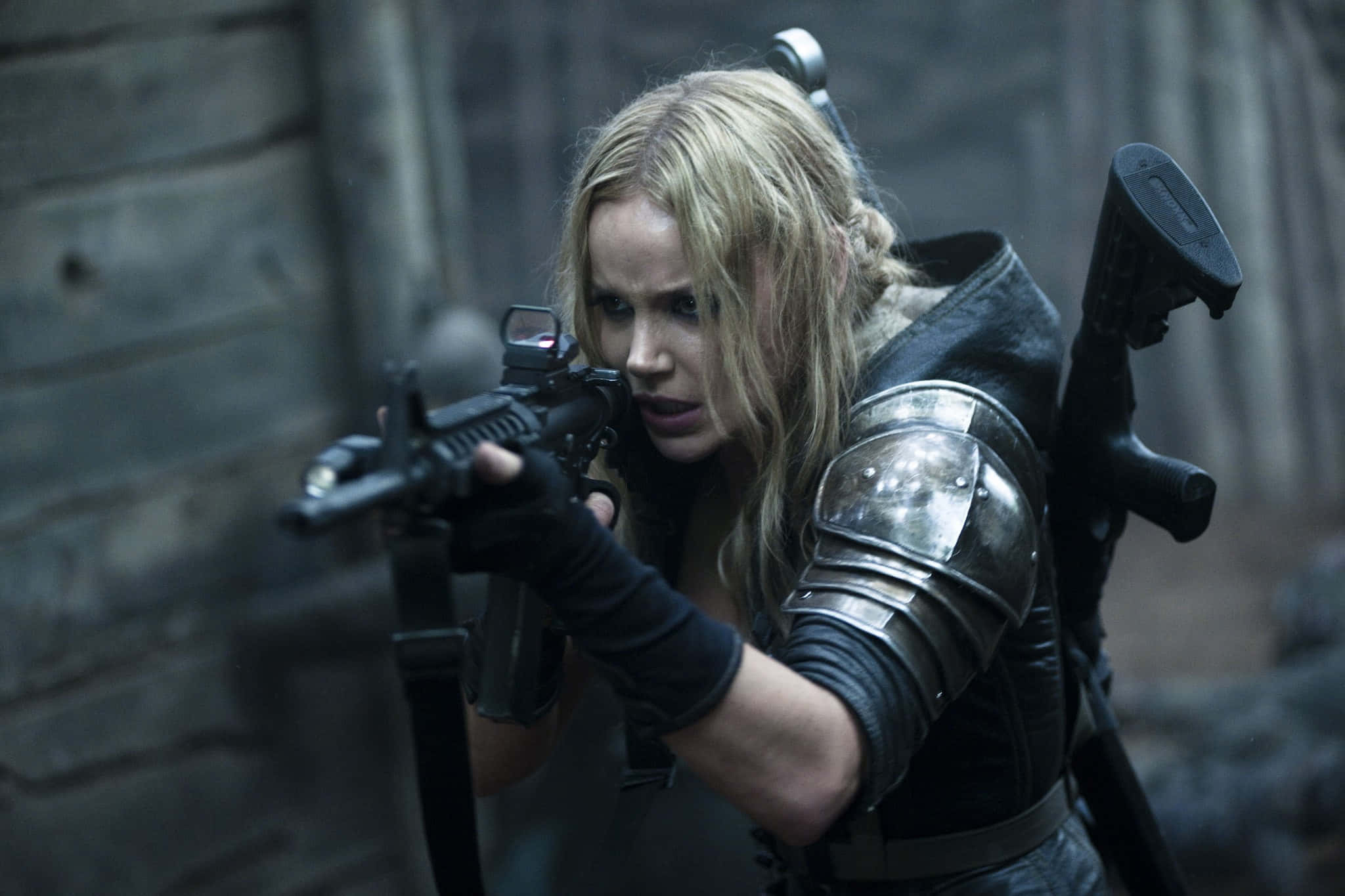 Abbie Cornish 桌布 壁紙 — 一位金髮女子身穿盔甲，在粗糙的石牆背景下，專注地瞄準步槍。