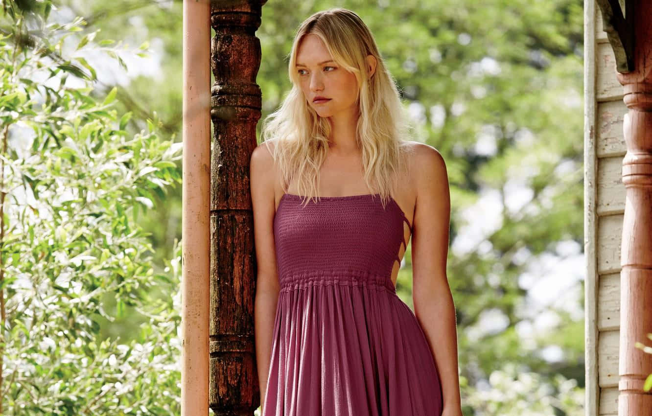 Gemma Ward 桌布 壁紙 — 一位金髮女子身穿紫色連衣裙，在斑駁的陽光下靠著木柱，周圍是茂密的綠色樹葉。