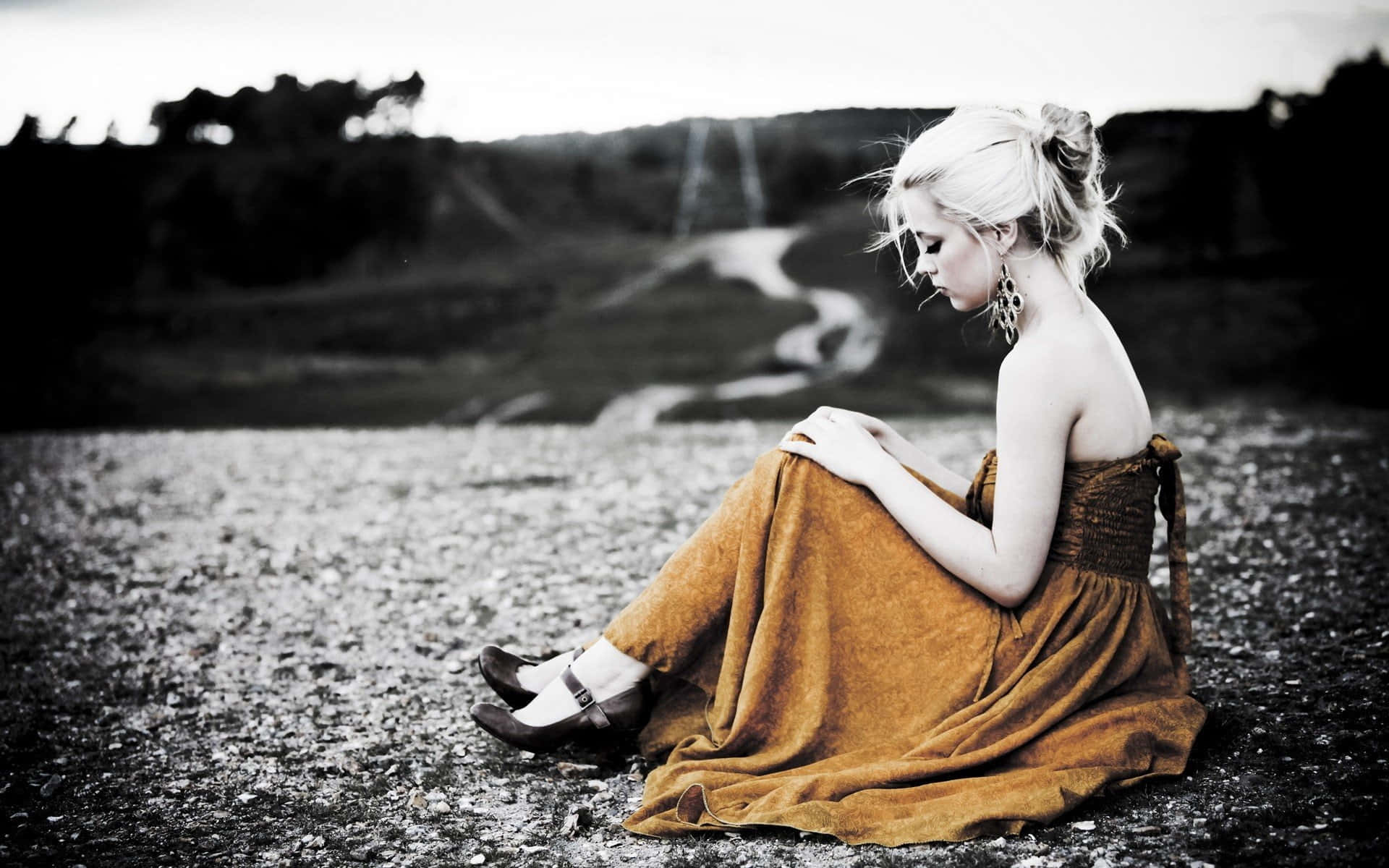 Alone 桌布 壁紙 — 一位金髮女子身穿芥末黃連衣裙，獨自坐在碎石地上，低頭凝視，一條蜿蜒的小徑在她身後逐漸消失在朦朧的山丘中。