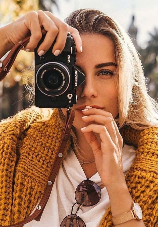 Holding Camera 桌布 壁紙 — 一位金髮女子身穿芥末黃針織開襟衫，手持黑色Olympus相機貼近眼睛，若有所思地凝視著觀者。