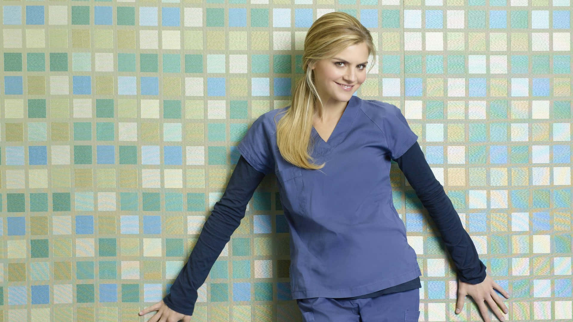 eliza coupe 桌布 壁紙 — 一位金髮女子身穿藍色手術服，靠著色彩繽紛的瓷磚牆，露出溫暖的笑容。