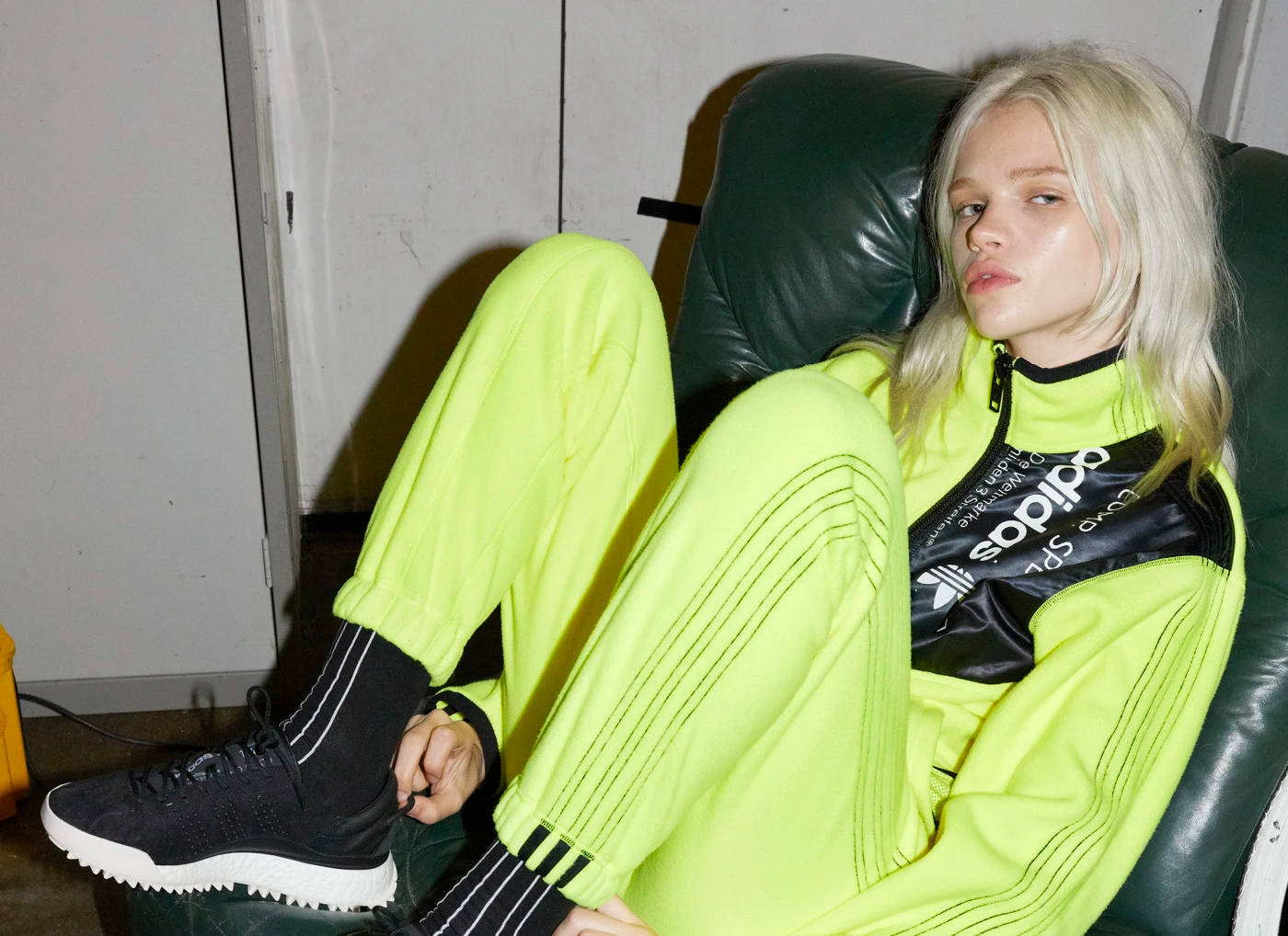 Alexander Wang 桌布 壁紙 — 一位金髮女子身穿螢光黃色Adidas運動服，在燈光昏暗的房間裡，隨意地躺在一張深綠色皮革椅上。