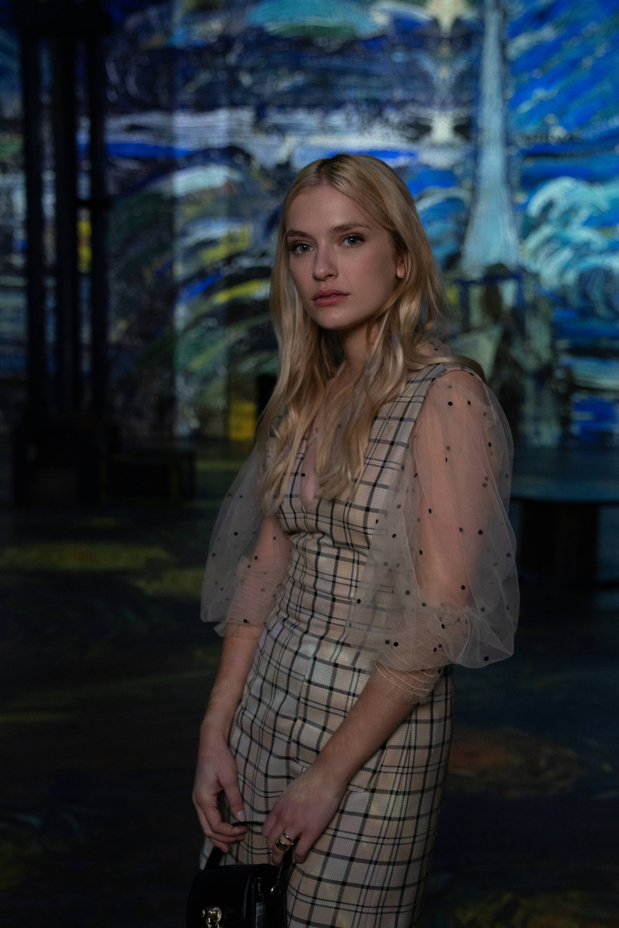 Emily In Paris 桌布 壁紙 — 一位金髮女子身穿透明袖的格子裙，站在昏暗畫廊中投影的梵谷藝術作品前。
