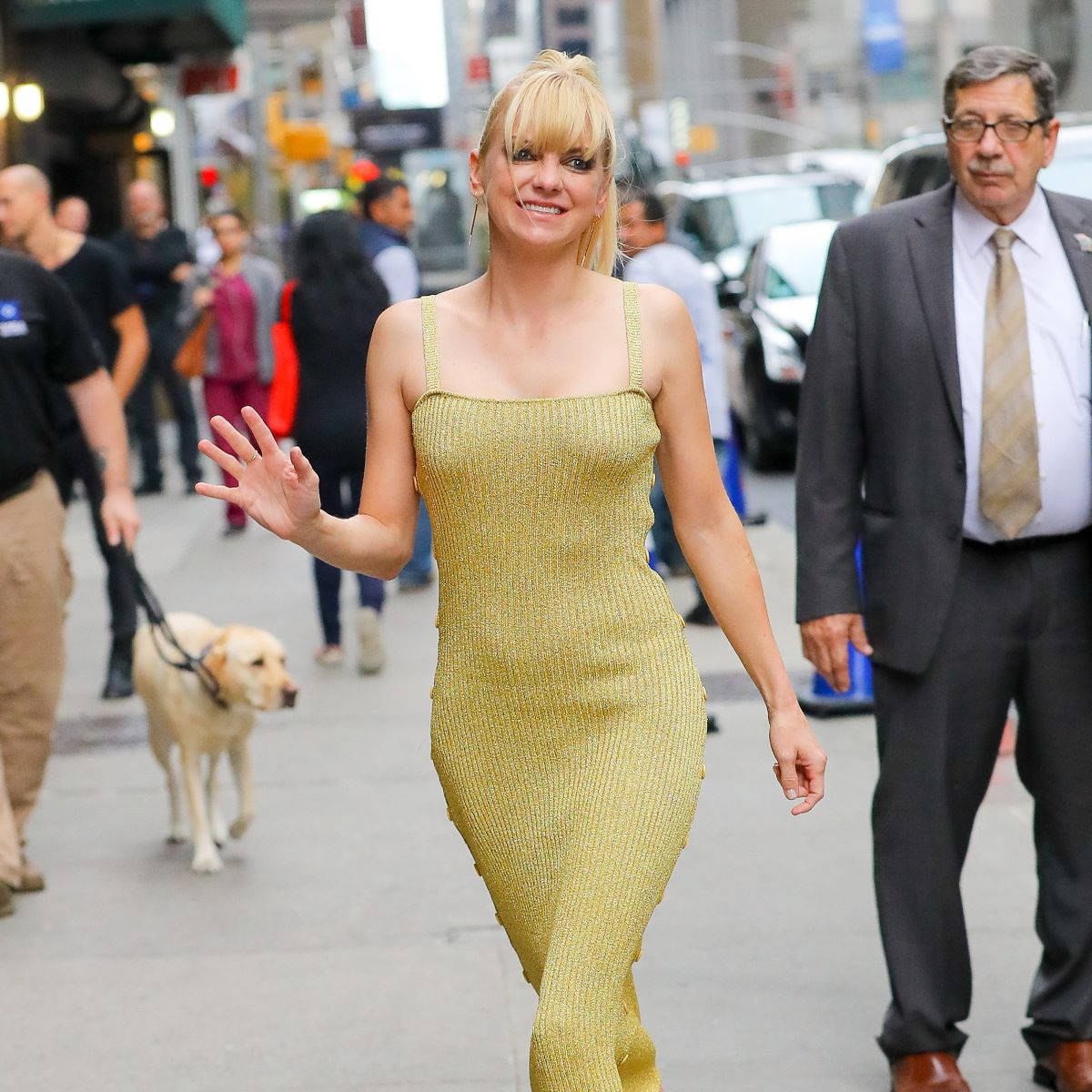 Celebrity 桌布 壁紙 — 一位金髮女子身穿閃閃發光的金色連衣裙，在繁忙的城市人行道上遛著一隻黃色拉布拉多犬，並愉快地揮手。