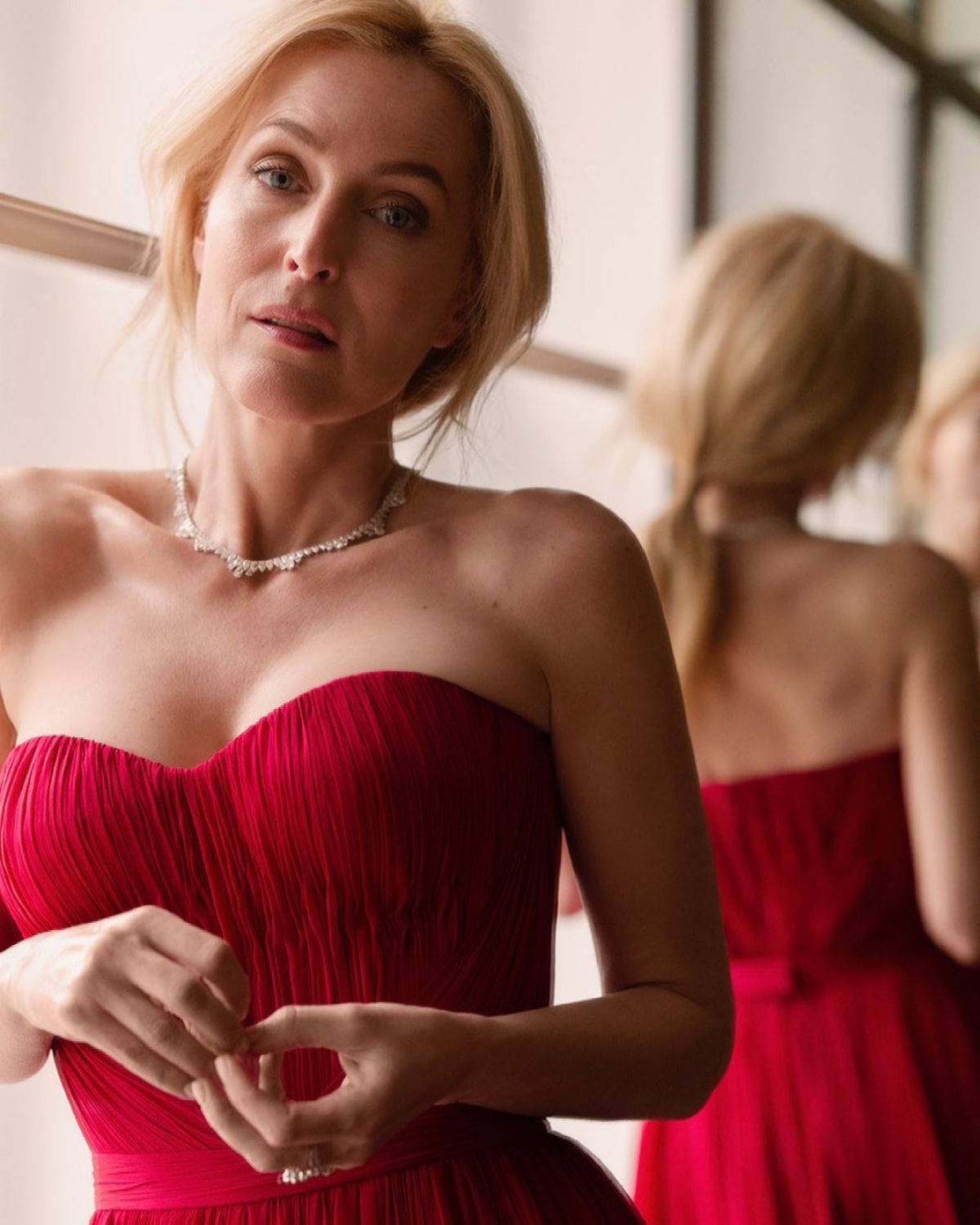 gillian anderson 桌布 壁紙 — 一位金髮女子身穿鮮紅色無肩帶禮服，一邊調整她的鑽石項鍊，一邊若有所思地看著鏡中的自己。