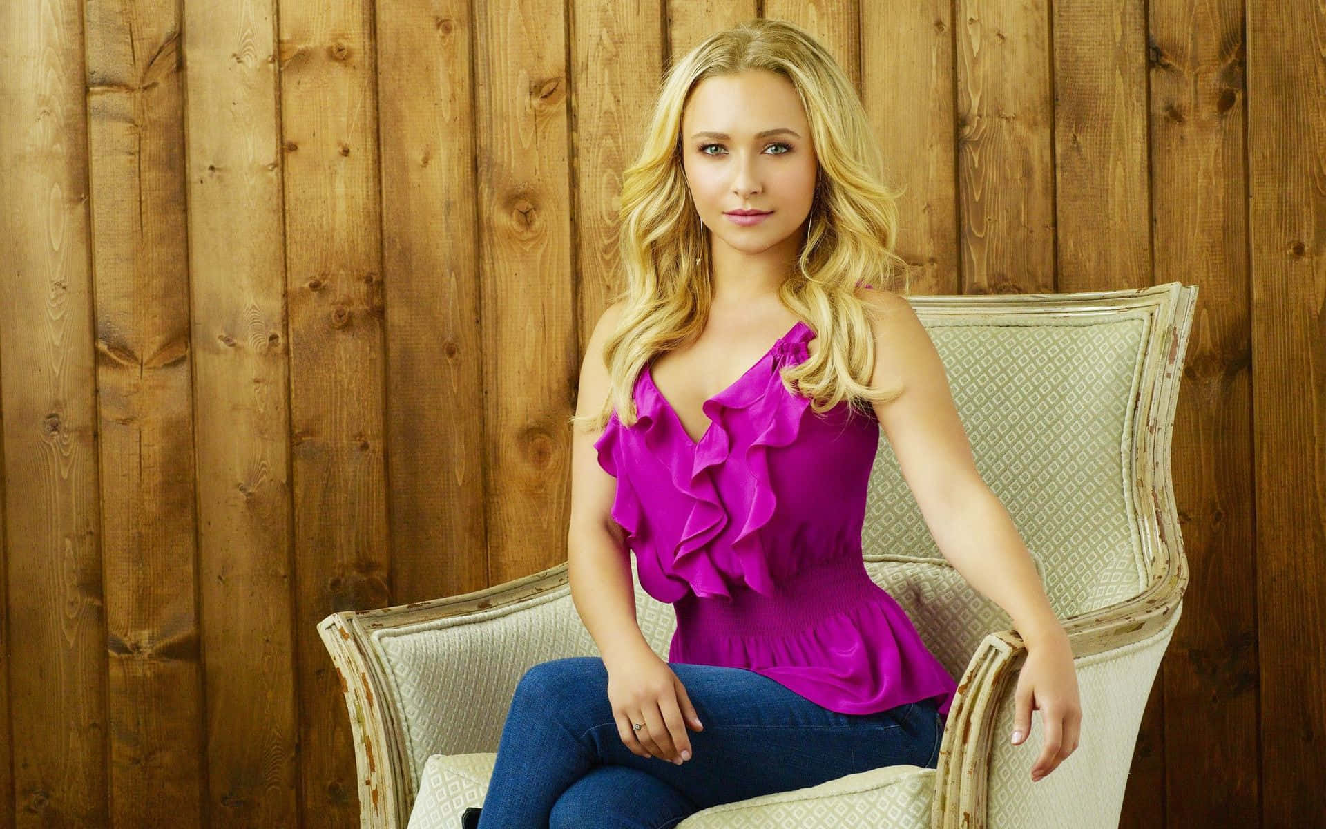 hayden panettiere 桌布 壁紙 — 一位金髮女子身穿鮮豔的紫紅色荷葉邊上衣，端坐在一張裝飾華麗的奶油色椅子上，背景是質樸的木牆。