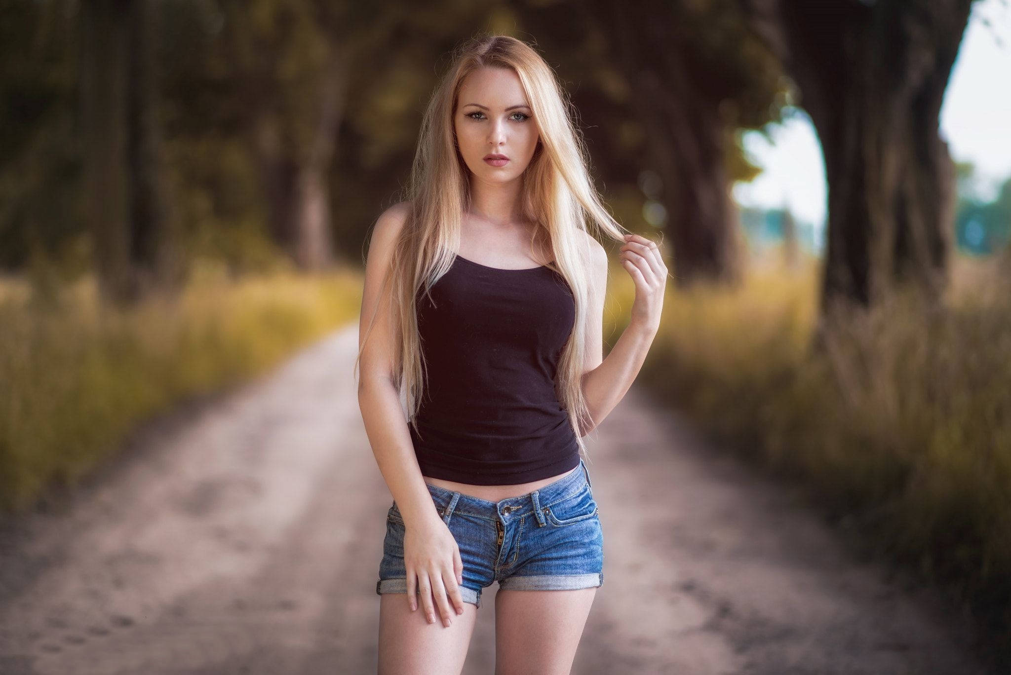 hair 桌布 壁紙 — 一位金髮女子身穿黑色上衣和牛仔短褲，自信地站在一條陽光普照、兩旁種滿高樹的泥土小徑上。