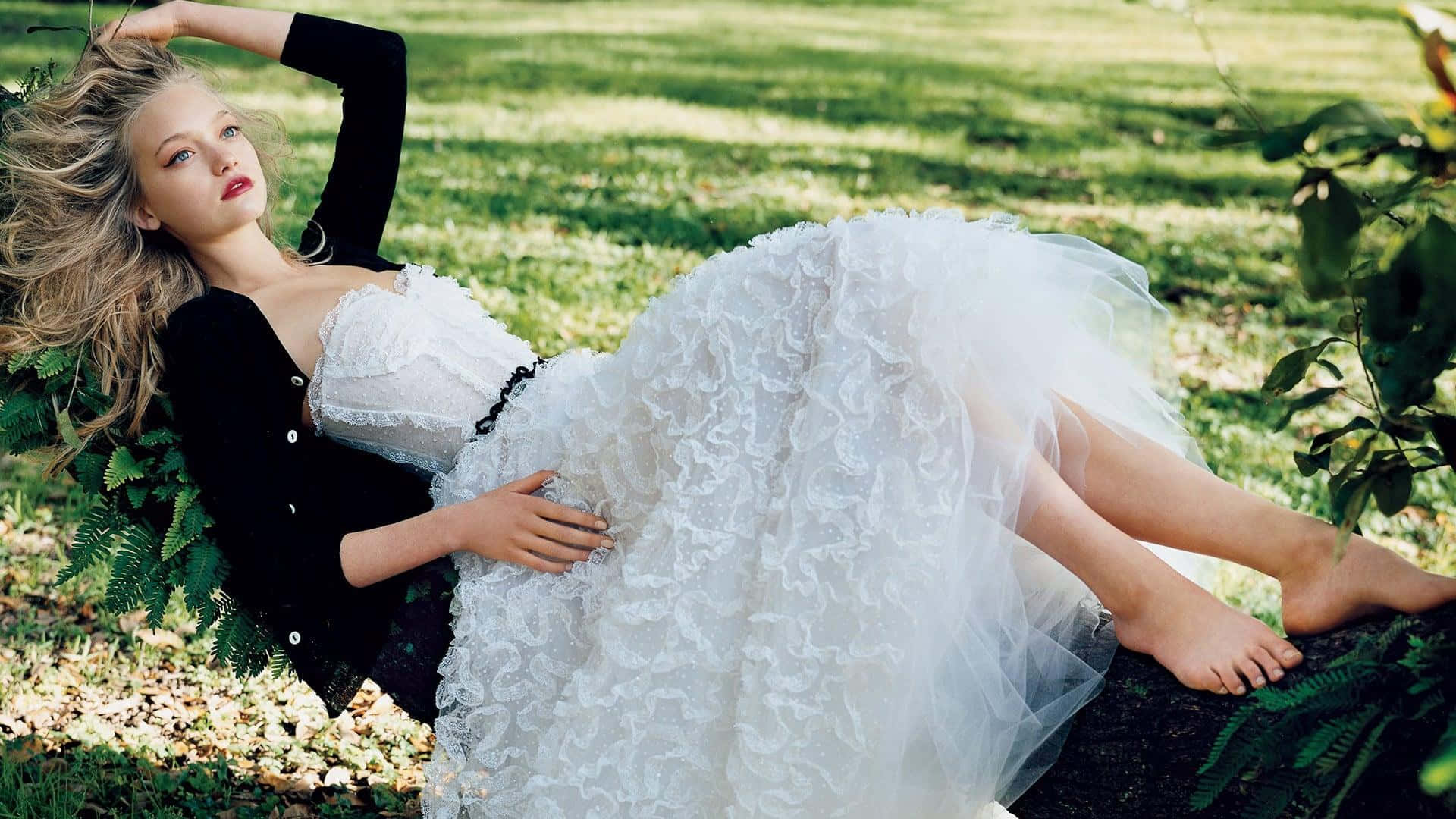 Gemma Ward 桌布 壁紙 — 一位金髮女子身穿黑色上衣和白色荷葉邊連衣裙，赤腳躺在陽光下蕨類植物間的草地上。