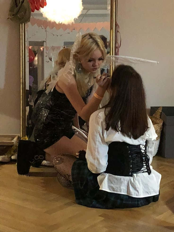 女同志 桌布 壁紙 — 一位金髮女子身穿黑色亮片服裝，帶有白色翅膀，跪著為另一位身穿白色襯衫和馬甲的女子化妝。