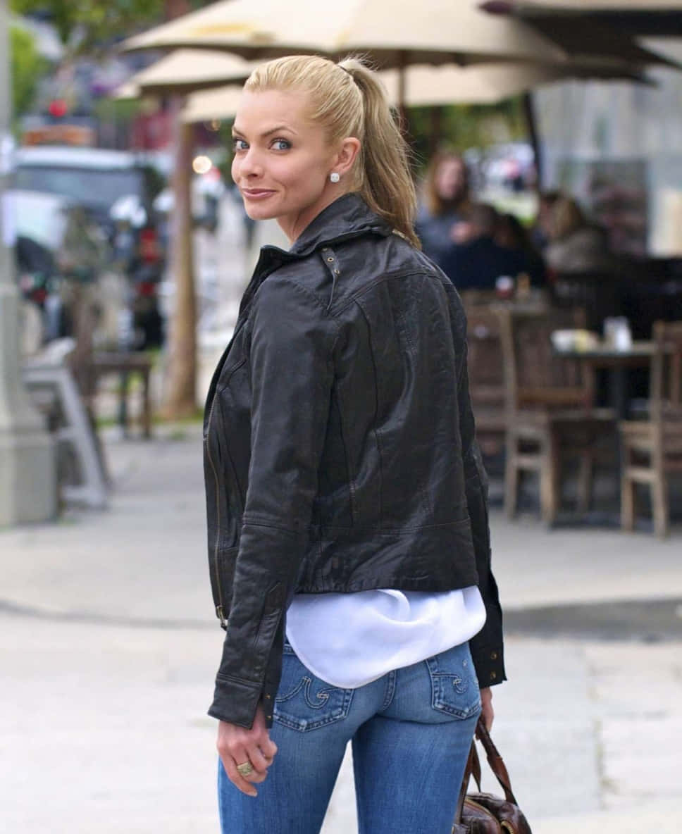Jaime Pressly 桌布 壁紙 — 一位金髮女子身穿黑色皮夾克和牛仔褲，在繁忙的城市人行道上轉身向鏡頭微笑。