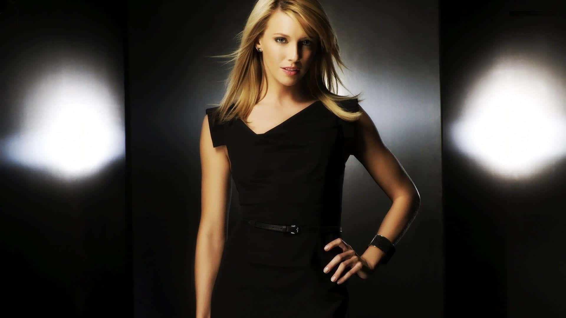 katie cassidy 桌布 壁紙 — 一位金髮女子身穿黑色連衣裙，在兩個明亮聚光燈照射的黑暗背景下自信地站立。