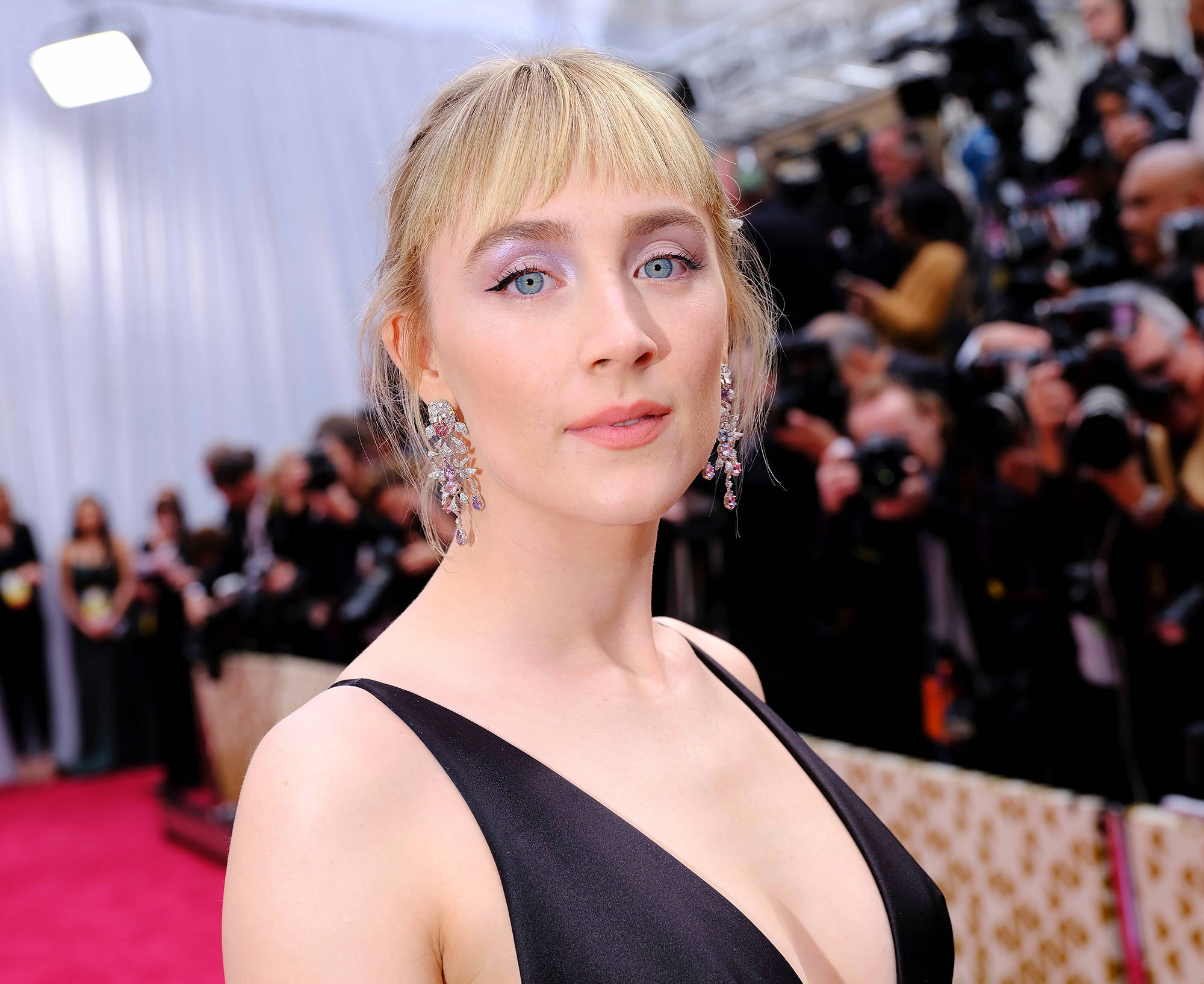 Saoirse Ronan 桌布 壁紙 — 一位金髮女子身穿黑色連衣裙和閃亮耳環，在紅地毯上擺姿勢，身後有攝影師。