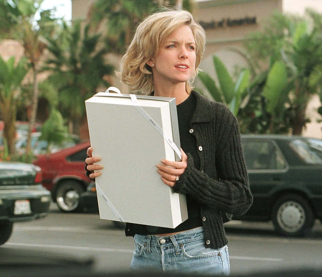 Courtney Thorne Smith 桌布 壁紙 — 一位金髮女子身穿黑色開襟衫和牛仔褲，站在陽光普照的街道上，手持一個大銀色禮物盒，周圍有棕櫚樹和汽車。