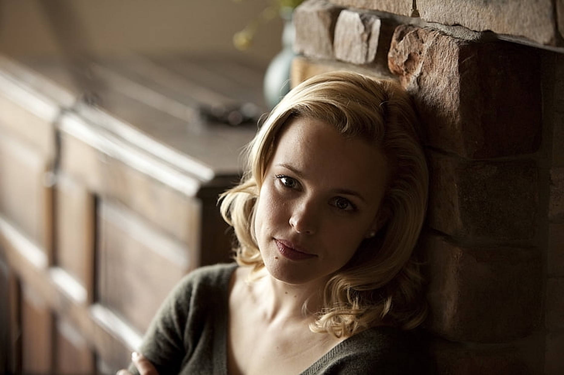 Rachel Mcadams 桌布 壁紙 — 一位金髮女子靠在質樸的磚砌壁爐旁，柔和溫暖的燈光突顯她沉思的表情。