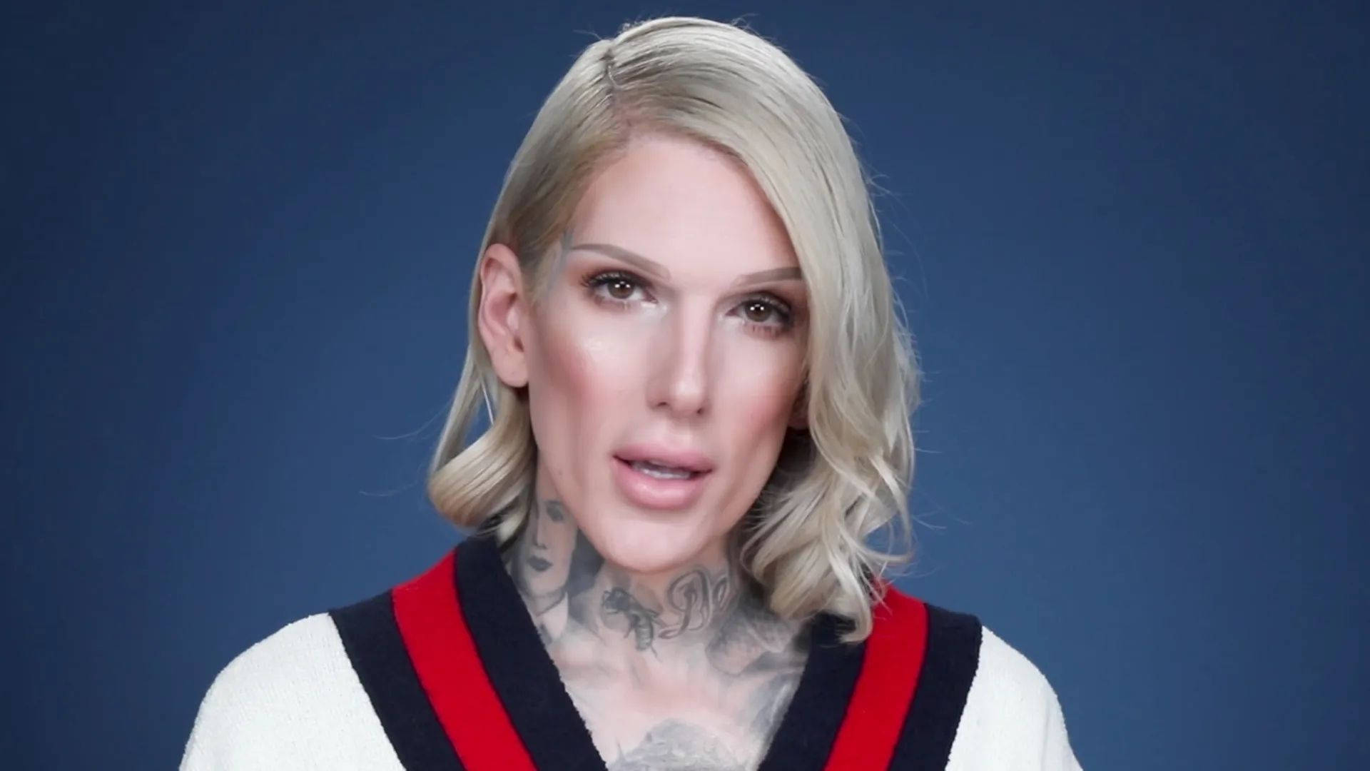 Jeffree Star 桌布 壁紙 — 一位金髮女子頸部有刺青，身穿帶有紅色和黑色條紋的白色毛衣，背景為純藍色。