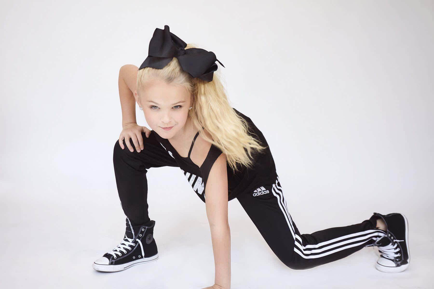 JoJo Siwa 桌布 壁紙 — 一位金髮女孩穿著黑色Adidas運動服和超大蝴蝶結，在純白色背景前擺出俏皮的蹲姿。