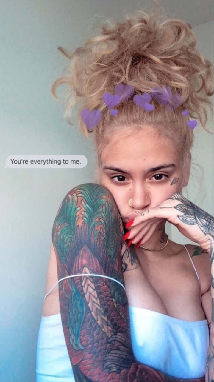 kehlani 桌布 壁紙 — 一位金髮女性，頭髮紮成凌亂的髮髻，飾有紫色心形，將有刺青的手臂靠近臉龐，靠在素色牆上。