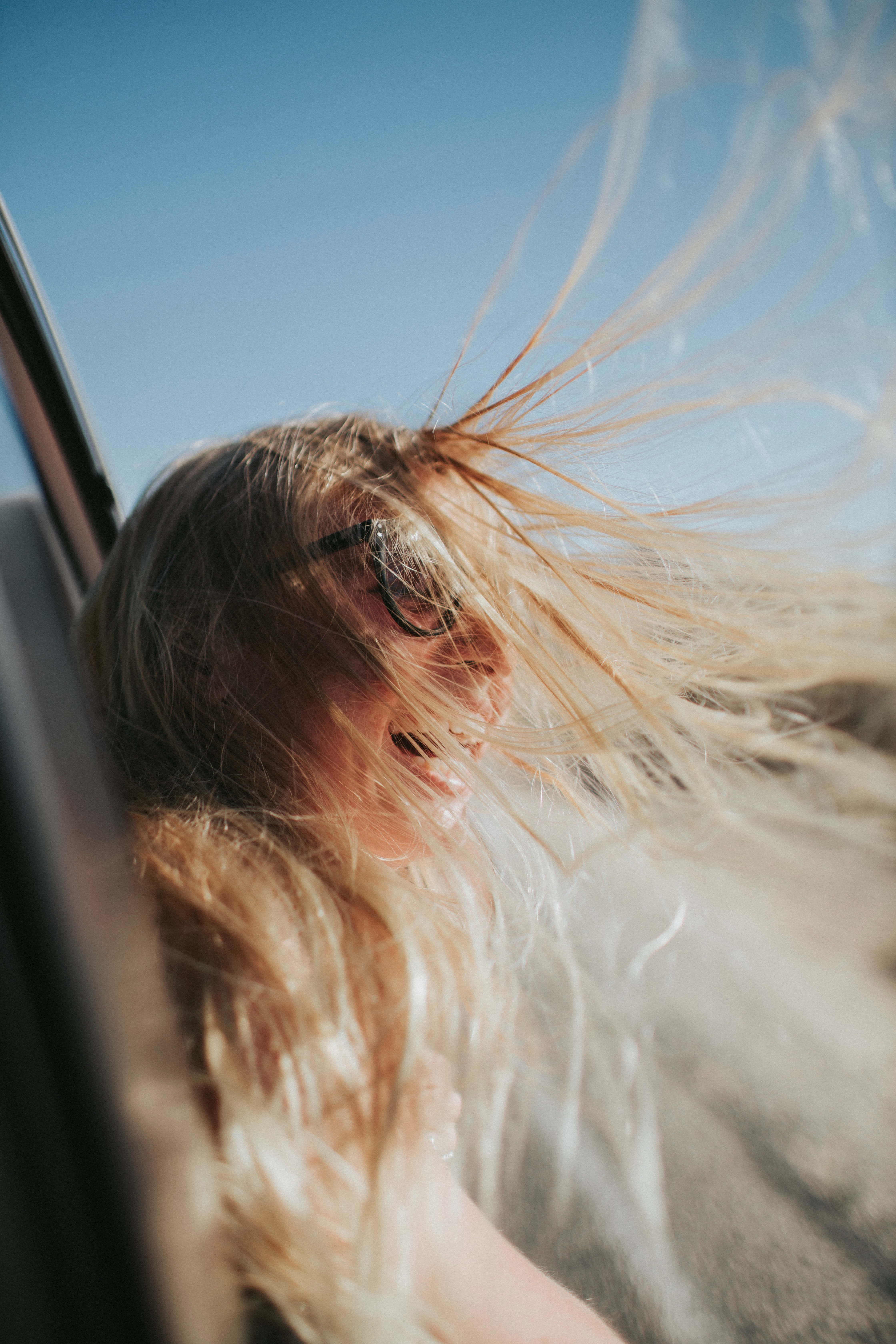 Woman 桌布 壁紙 — 一位金髮女性在晴朗藍天下從車窗探出頭，頭髮在風中狂亂飛舞，大笑著。