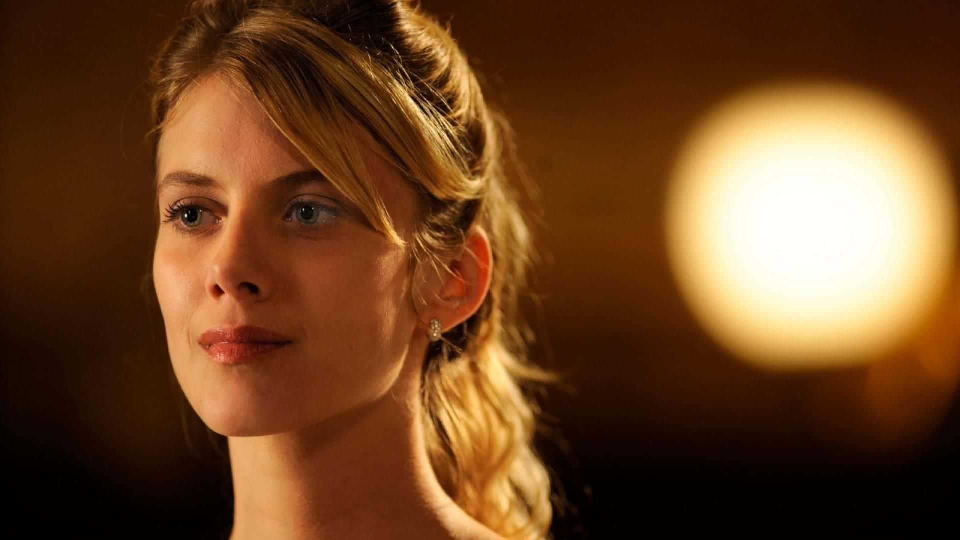 Mélanie Laurent 桌布 壁紙 — 一位金髮女性在柔和模糊的背景前，於溫暖的金色光芒中若有所思地凝視。