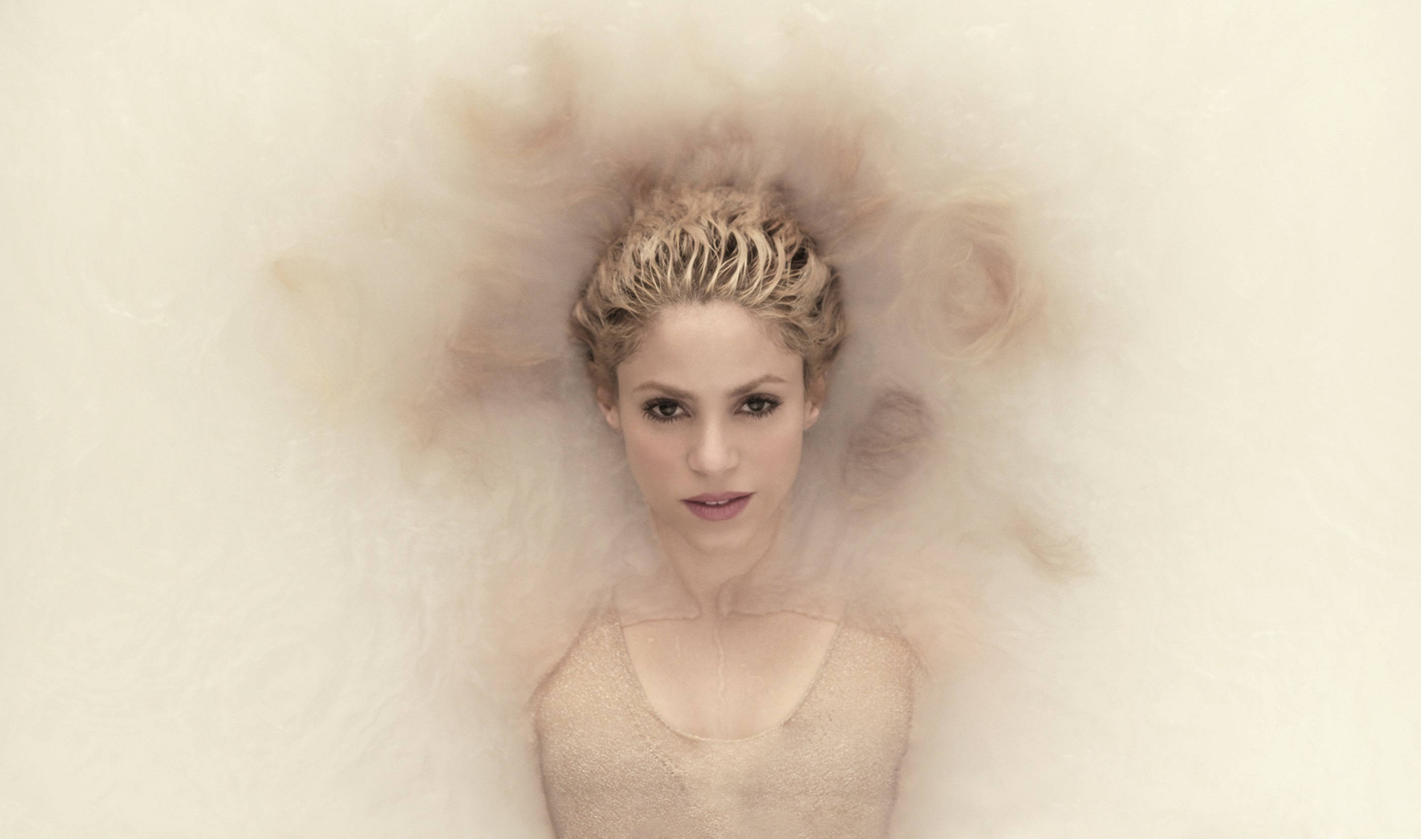 Shakira 桌布 壁紙 — 一位金髮女性在淡色背景前，於柔軟的米色粉末雲中寧靜地漂浮。