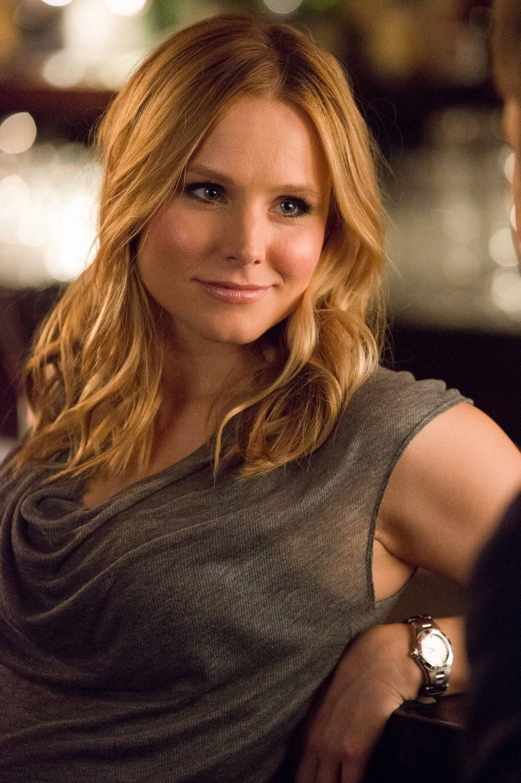 kristen bell 桌布 壁紙 — 一位金髮女性在溫暖燈光的室內環境中，穿著灰色上衣和手錶，溫柔地微笑。