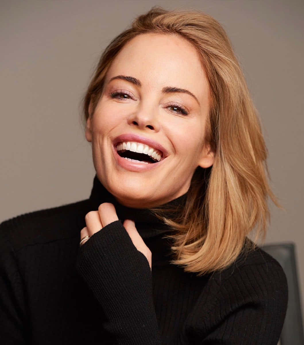 Chandra West 桌布 壁紙 — 一位金髮女性在灰色中性背景前穿著黑色高領毛衣，開懷大笑。