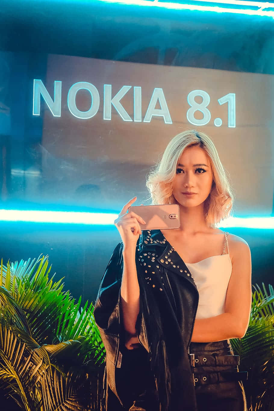 諾基亞 桌布 壁紙 — 一位金髮女性在霓虹燈下，於發光的Nokia 8.1標誌前手持電話，周圍環繞著熱帶植物。