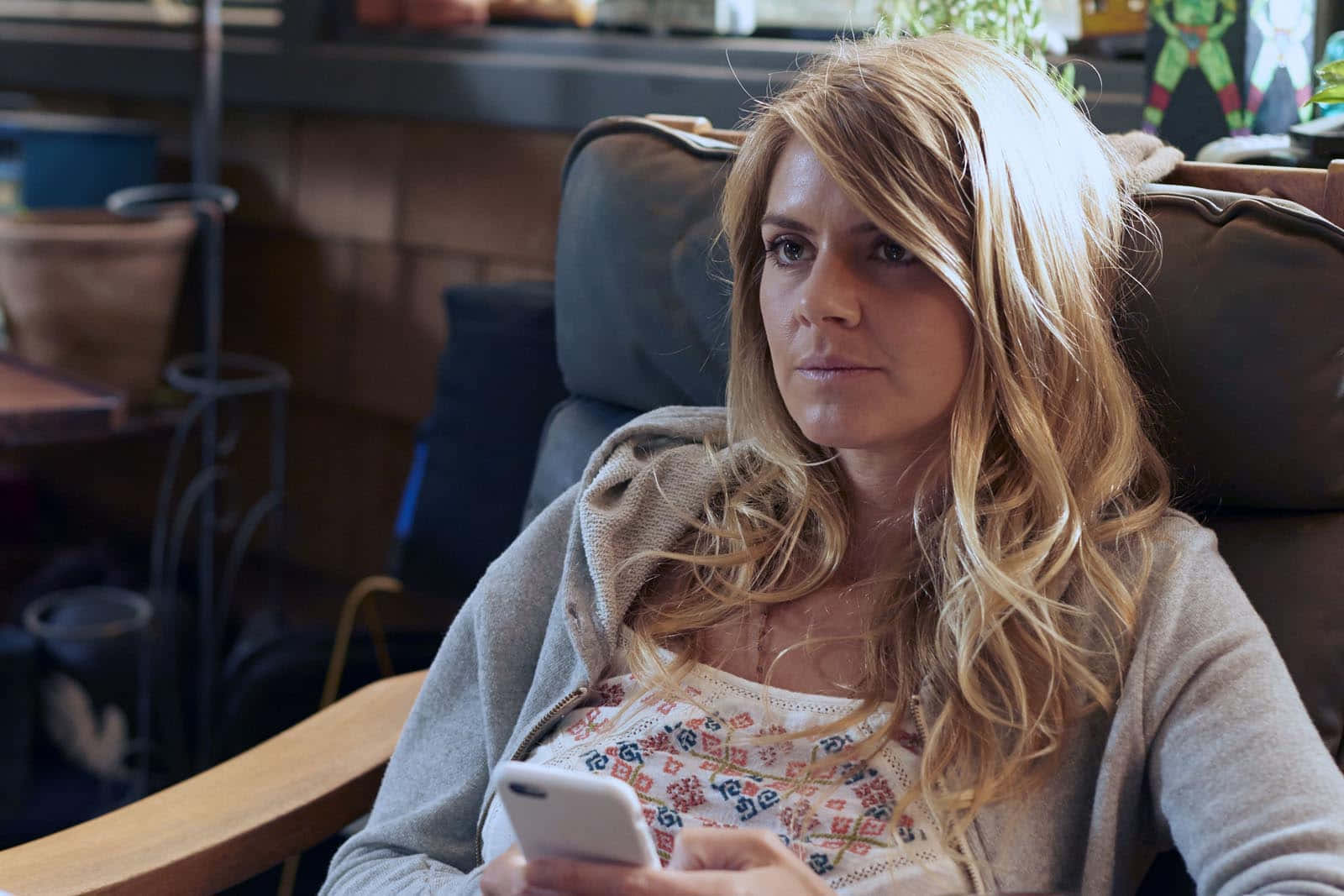 eliza coupe 桌布 壁紙 — 一位金髮女性坐在椅子上，手持電話，在舒適的室內環境中顯得若有所思。
