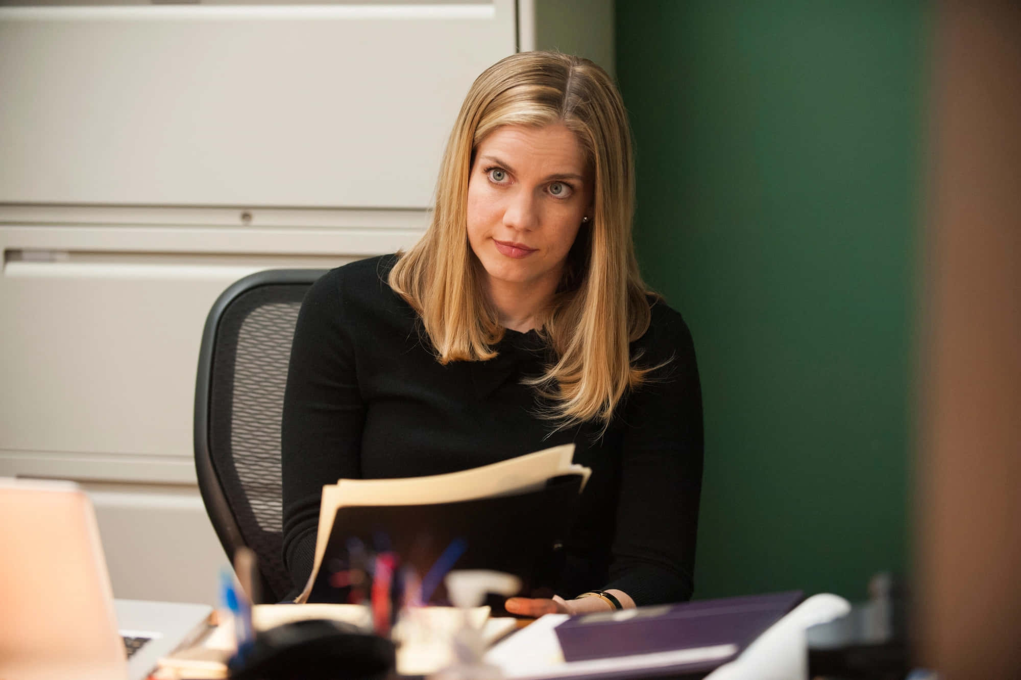 anna chlumsky 桌布 壁紙 — 一位金髮女性坐在雜亂的辦公桌前，手持文件，專注地看著相機。
