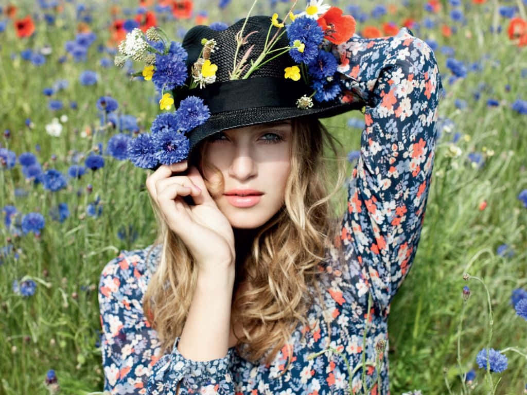 Spring Fashion 桌布 壁紙 — 一位金髮女性戴著一頂飾有藍色和黃色花朵的黑色帽子，站在一片生機勃勃的野花田中。